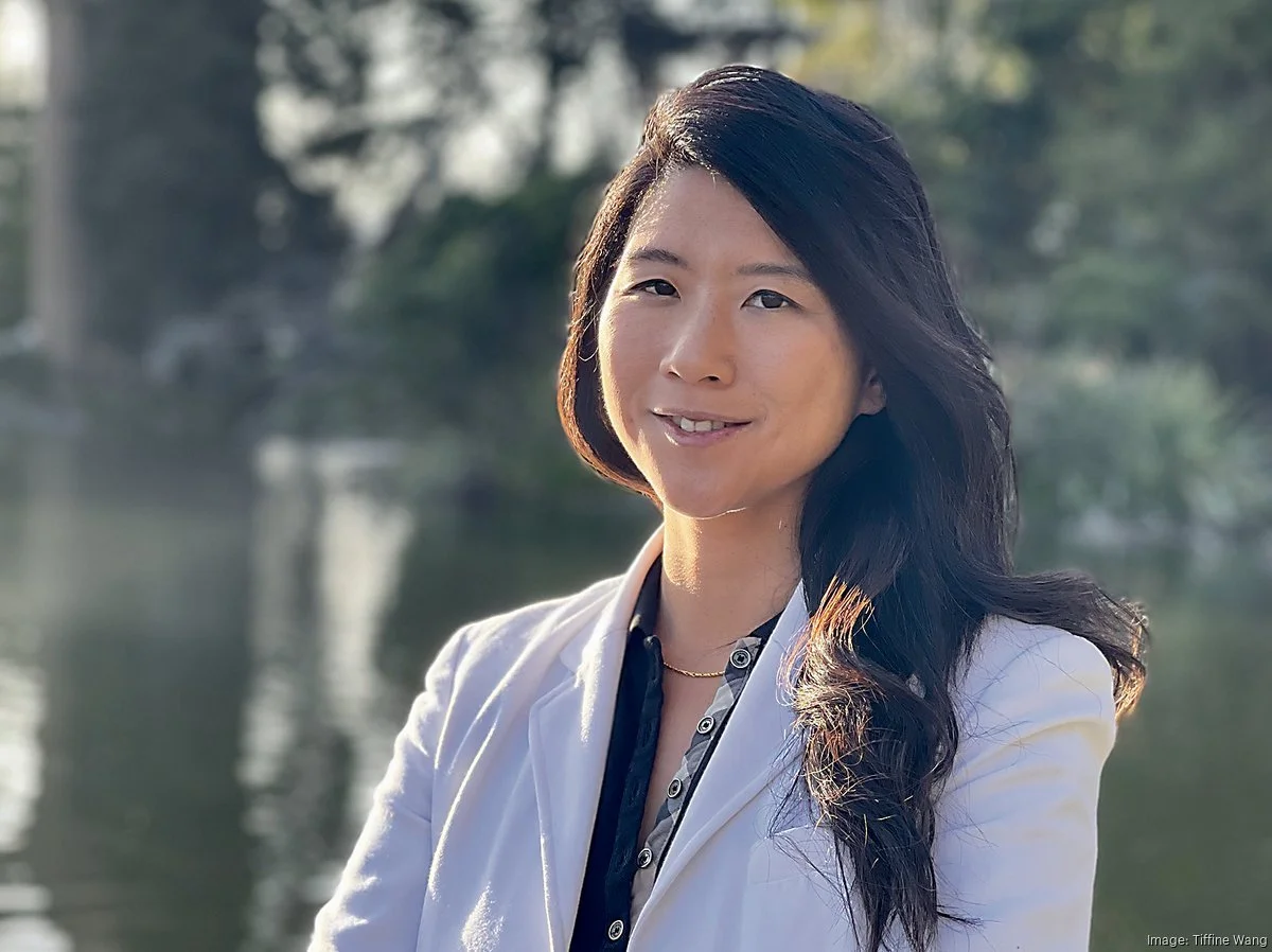 A woman with long black hair wearing a white blazer stands outdoors near water and trees.
