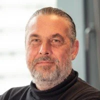 Close-up portrait of a middle-aged man with gray hair and beard, wearing a black shirt, indoors with a window in the background.