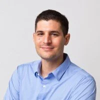 A young man with short dark hair, wearing a light blue collared shirt, smiling with arms crossed against a plain gray background.