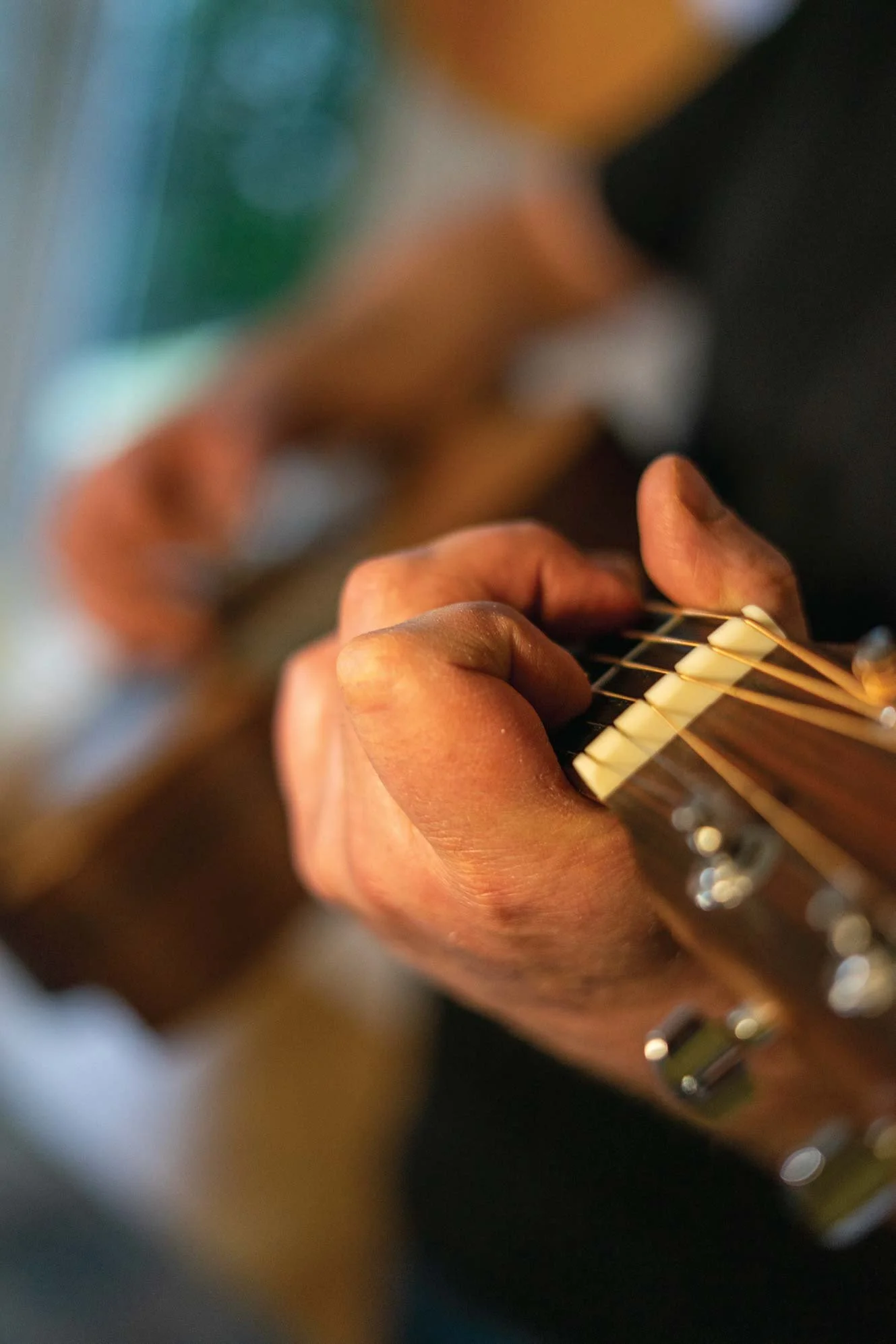 A person's hand is shown pressing down on the strings of an acoustic guitar, with the person's face blurred in the background.