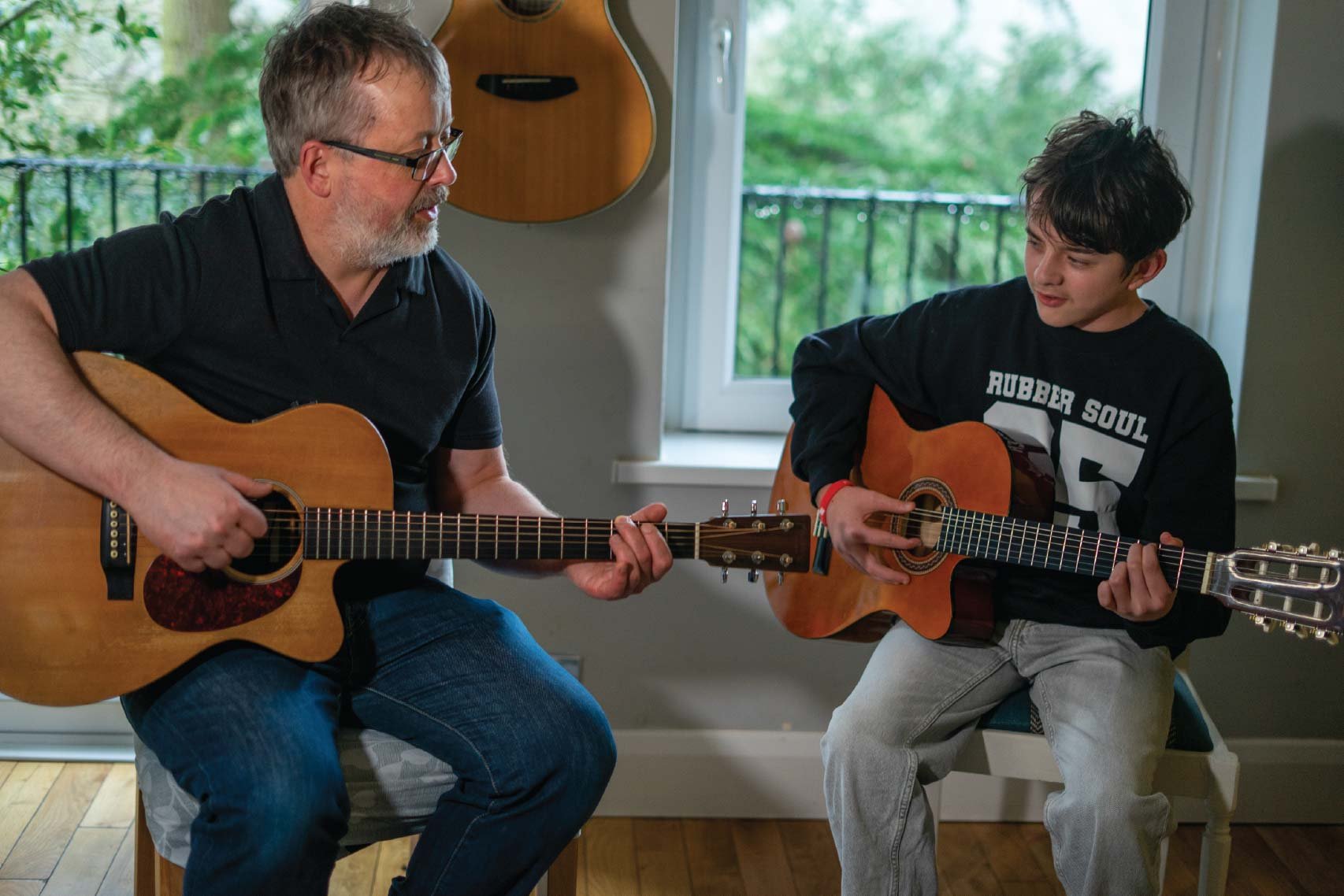 A man and a boy are playing acoustic guitars together in a room with a window, a guitar hanging on the wall, and natural light coming through the window.
