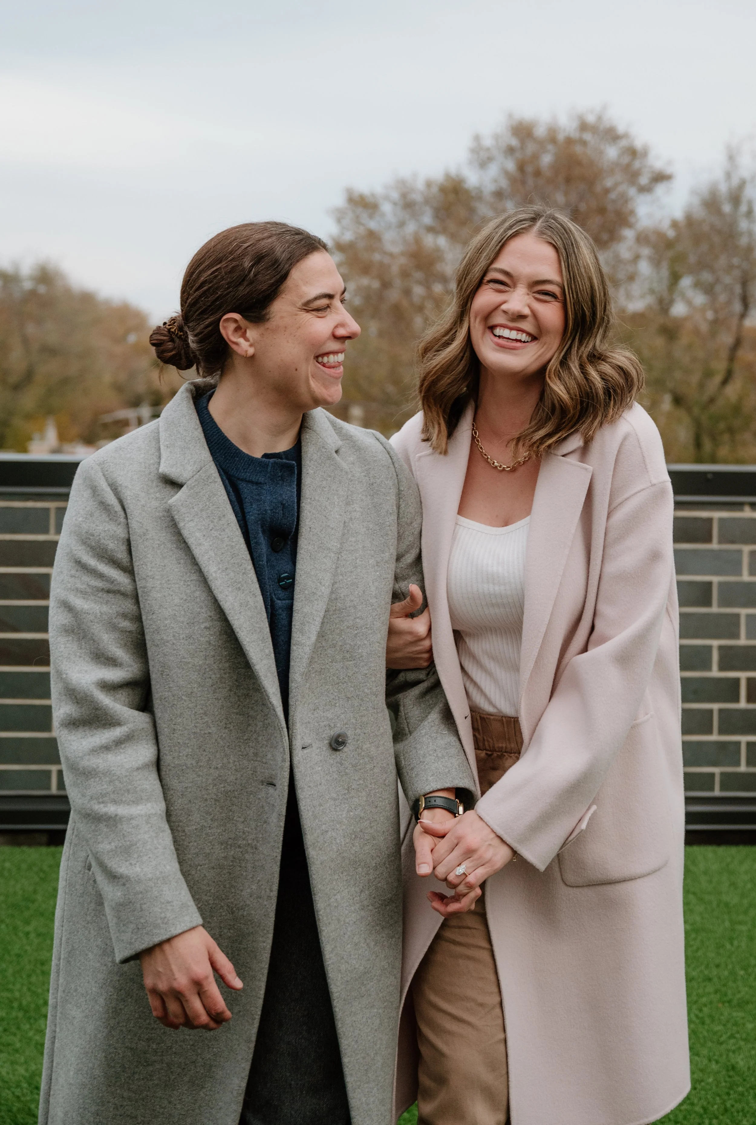 Two women standing outdoors, laughing and holding hands, autumn trees in the background.