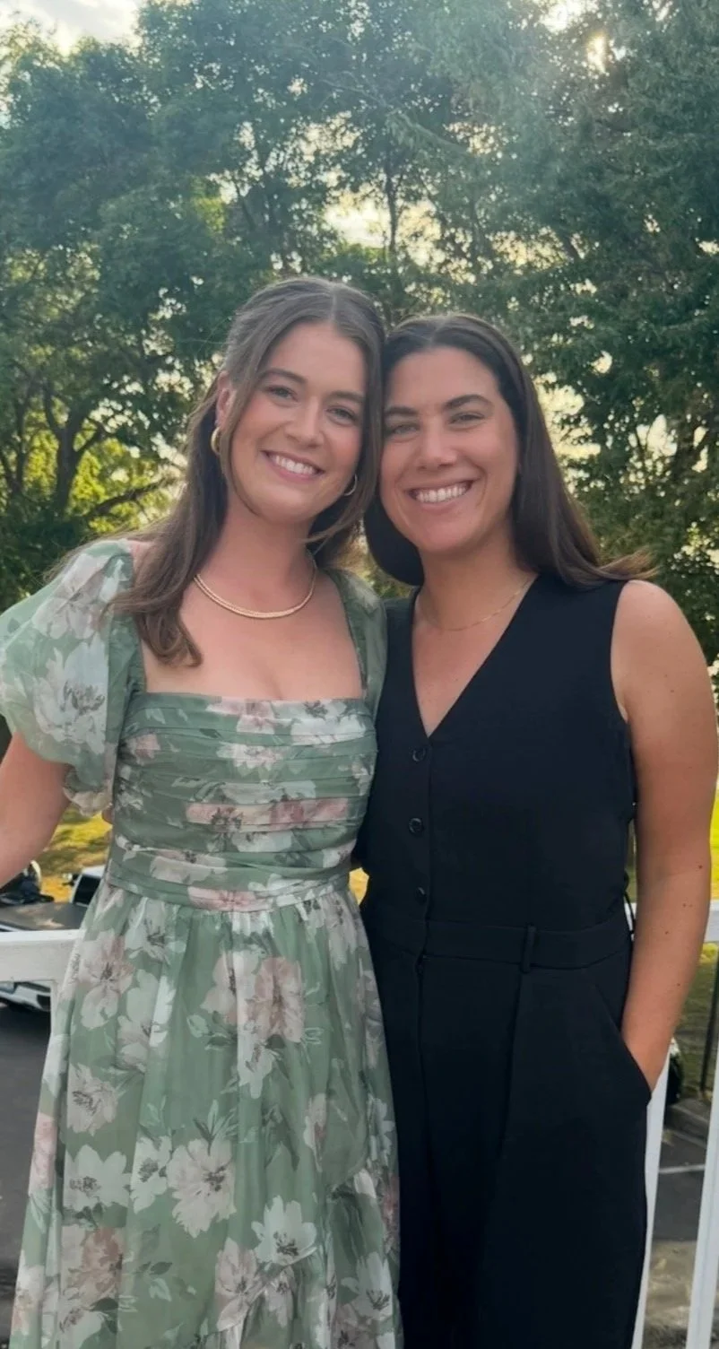 Two women smiling outdoors with trees and sunset in the background, one wearing a floral dress and the other in a black sleeveless top.