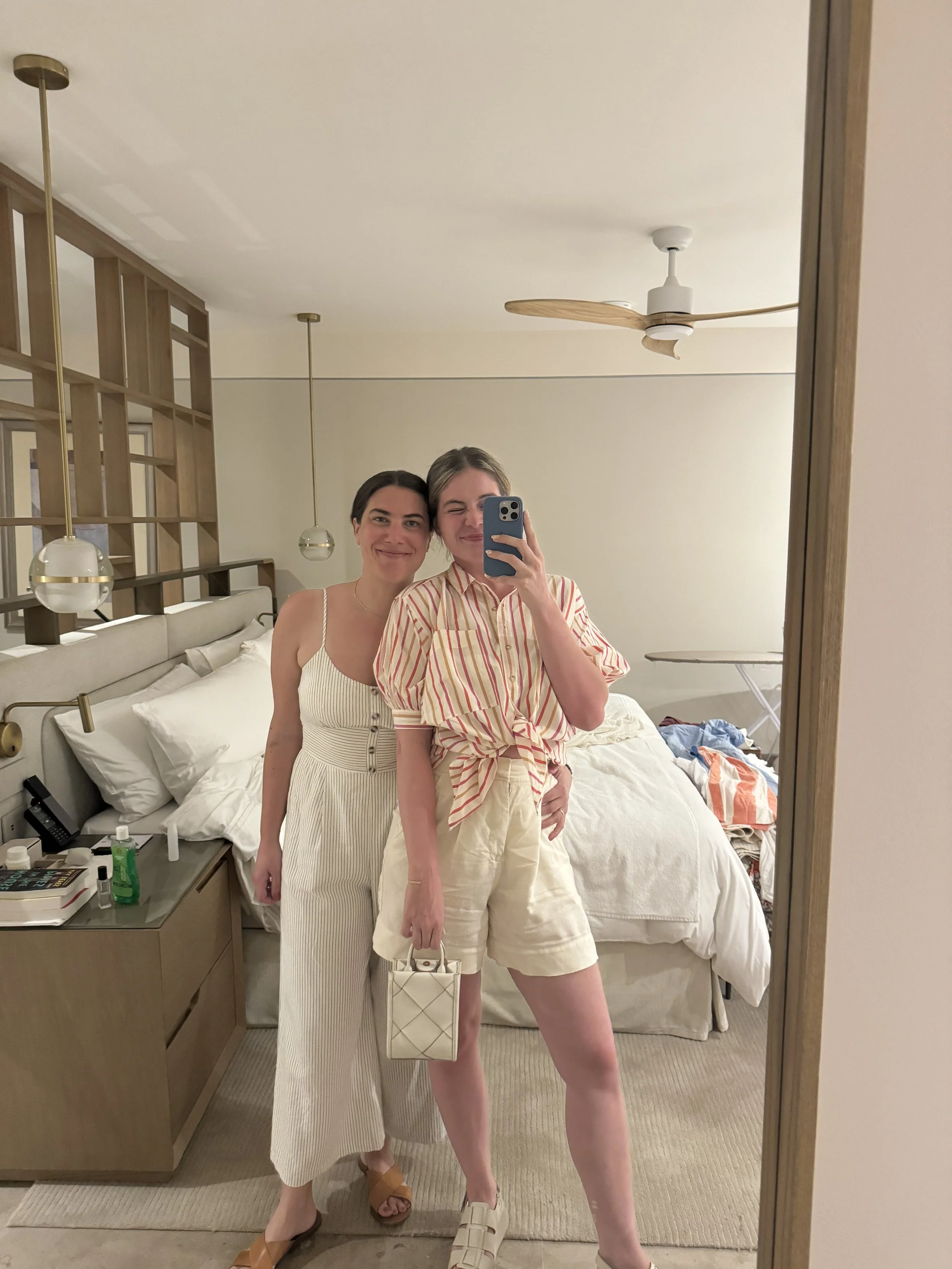 Two women taking a mirror selfie in a hotel room, smiling, with a bed, nightstand, and hanging lights in the background.