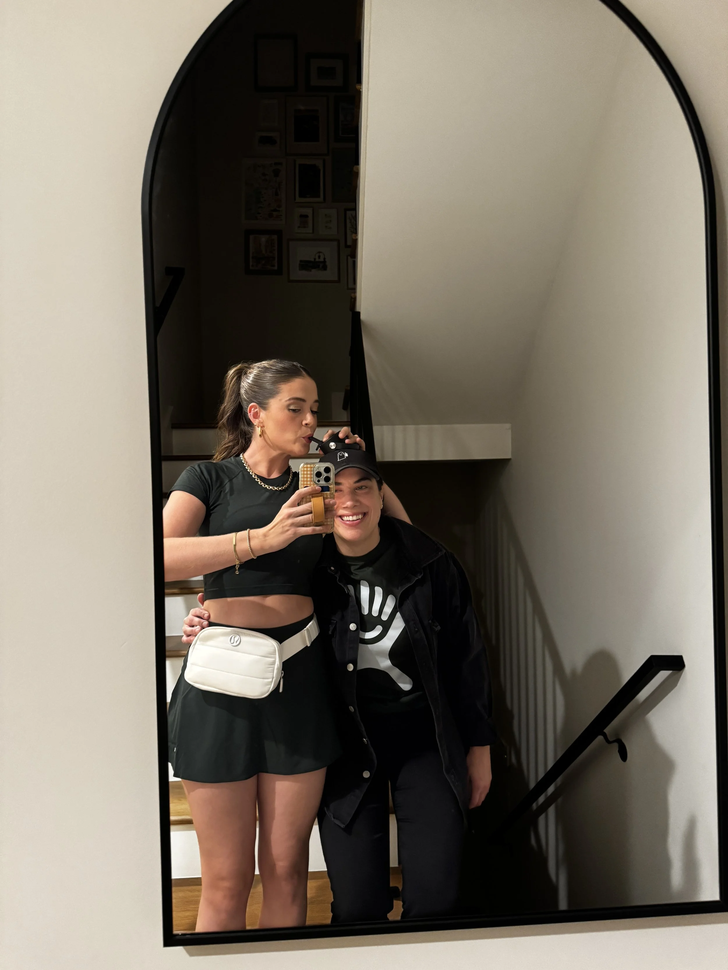 Two women taking a mirror selfie in a room with gallery wall art, one smiling and one making a playful face, both dressed casually.