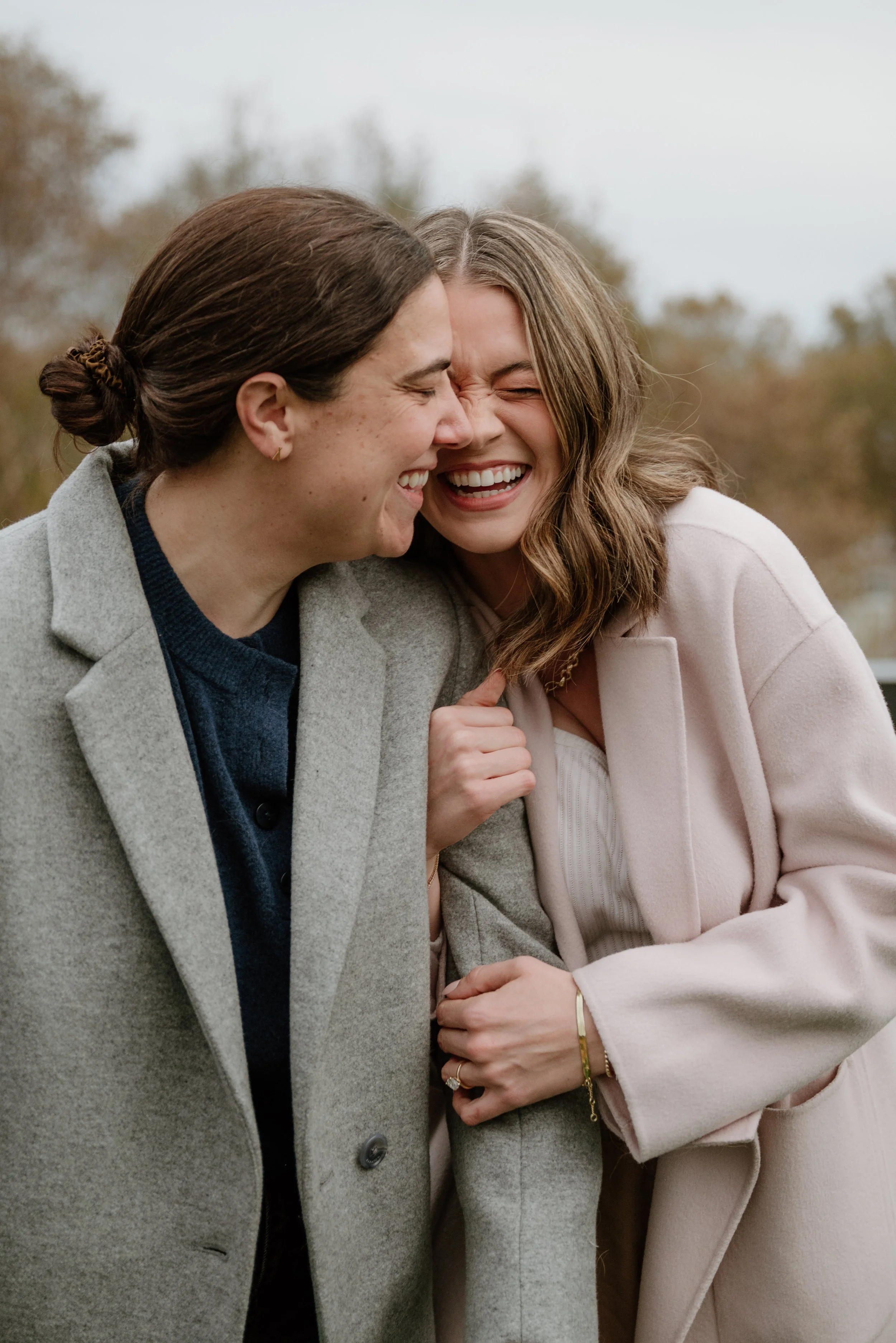 Two women are outside, smiling and laughing together, touching foreheads