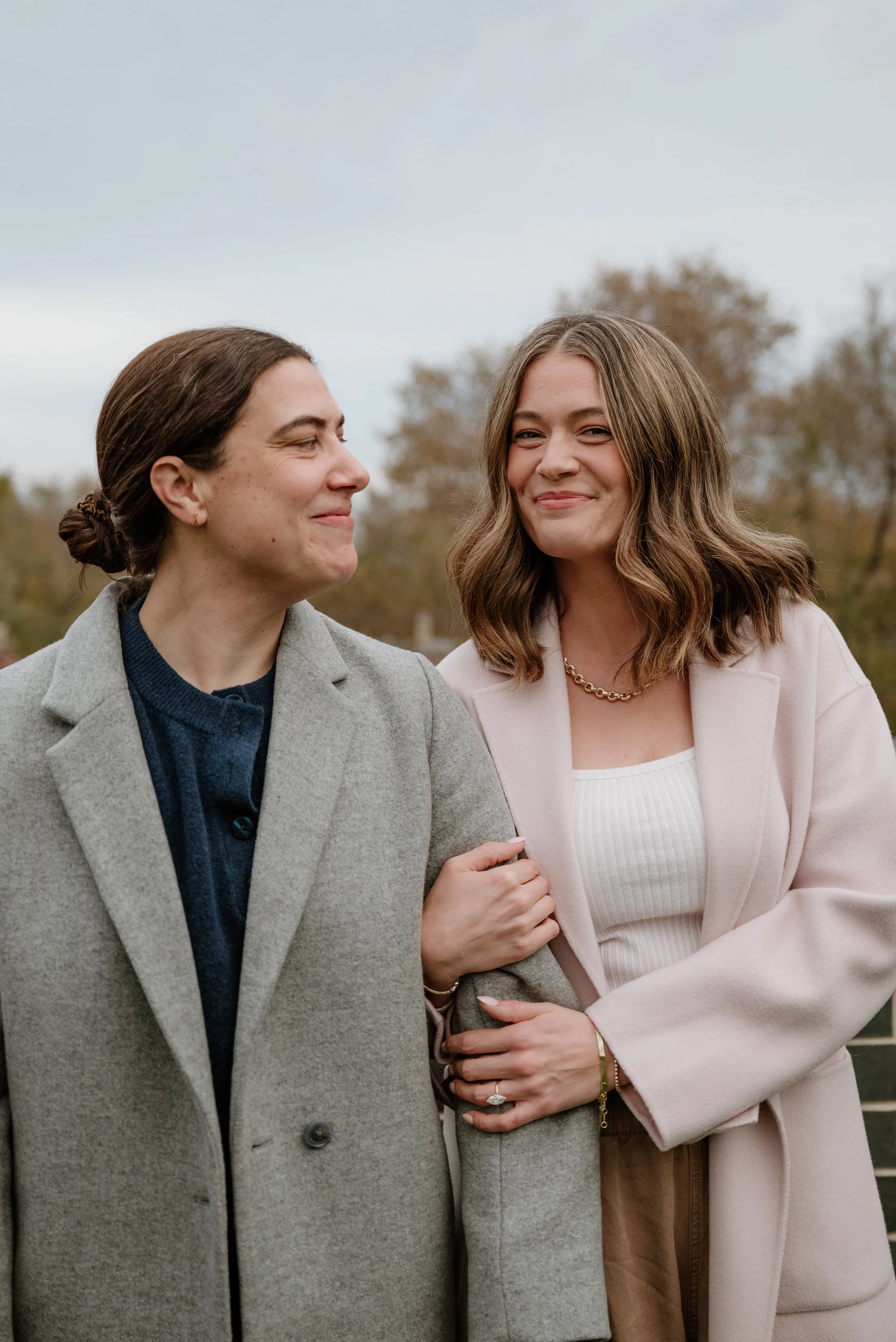 Two women standing close together outdoors, holding hands, smiling, with trees in the background, dressed in coats.