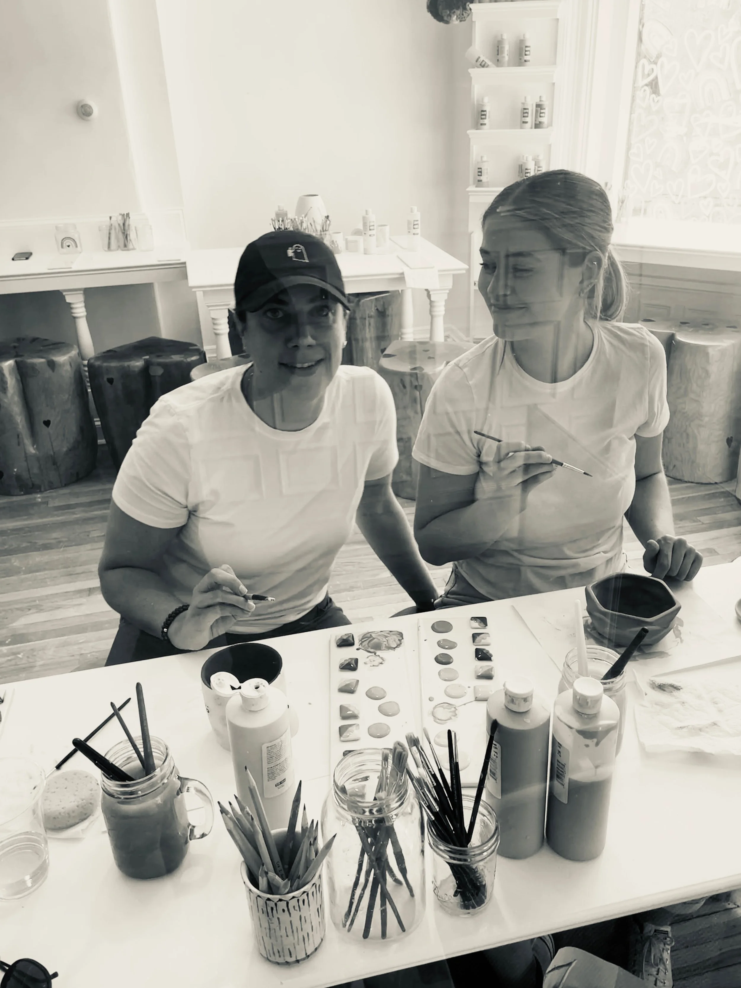 Two women sitting at a table with painting supplies. The table has jars of paint, brushes, and a palette. The women are smiling and wearing white t-shirts. The background features a bright room with a window and shelves with bottles.