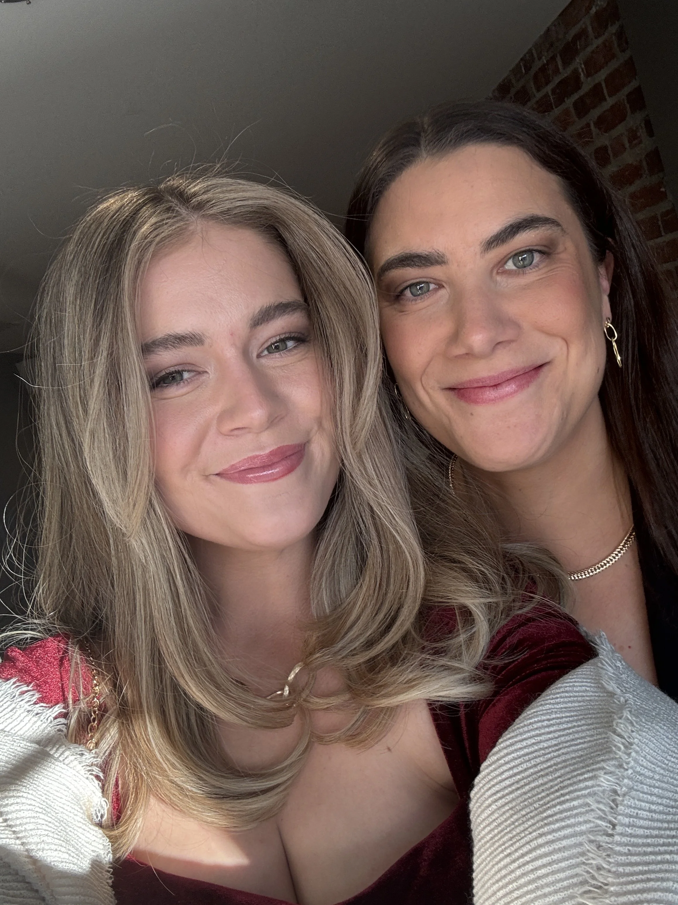 Close-up of two women smiling, with one woman having blonde hair and the other with dark hair, standing in front of a brick wall.