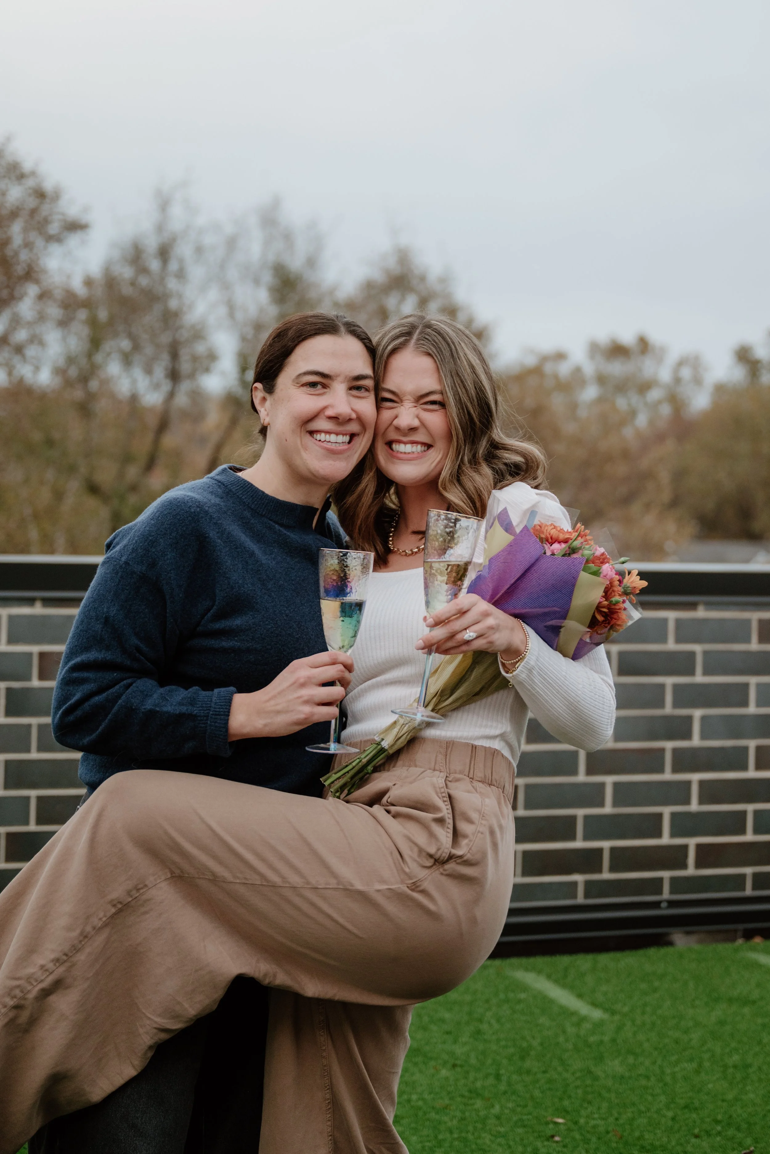 Two women smiling and celebrating, holding glasses of champagne and a bouquet, outdoors on a terrace with autumn-colored trees in the background.
