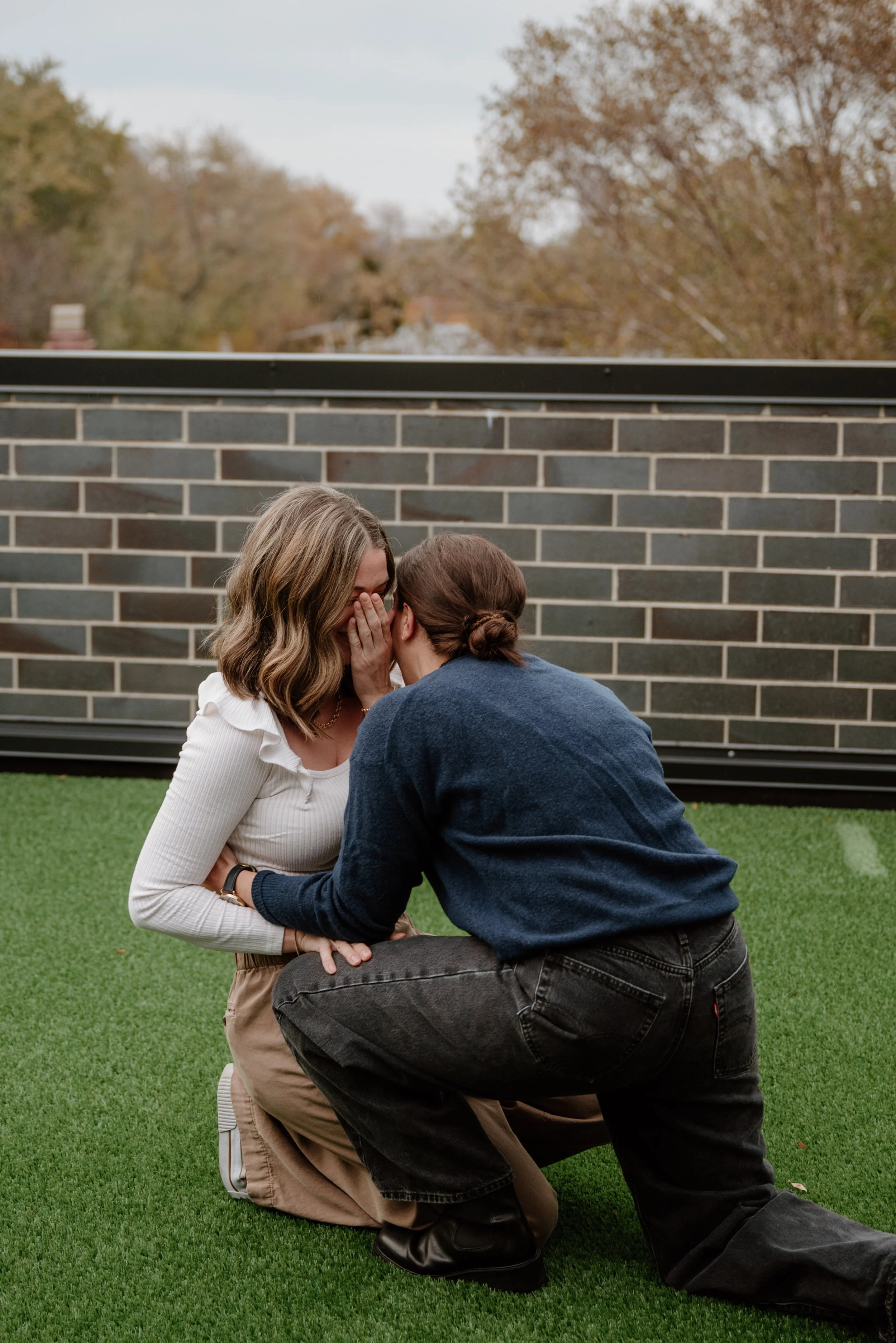 Two women stand close and whisper to each other, with one woman kneeling on the grass in front of the other. They are outdoors against a brick wall with trees and sky in the background.