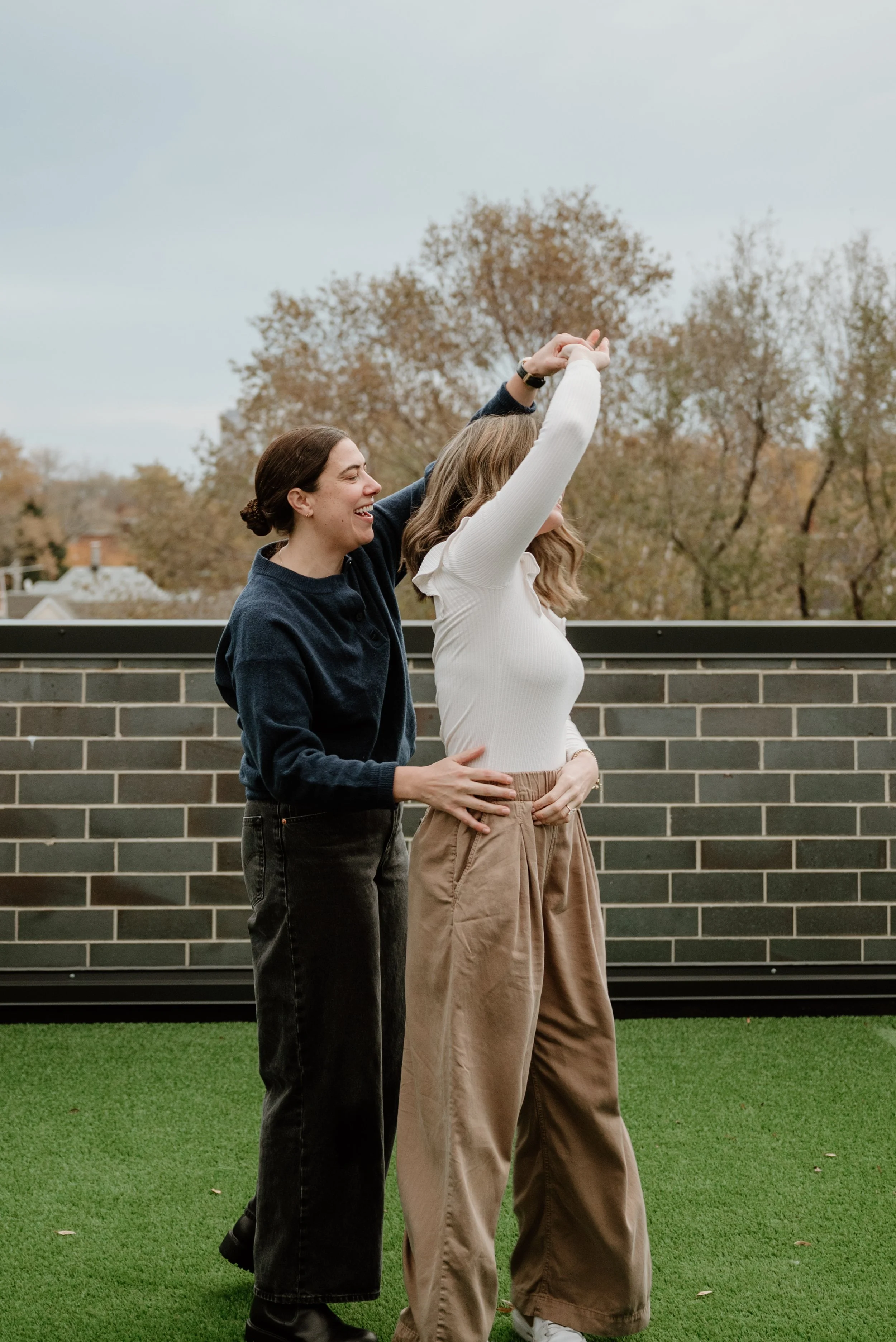 Two women dancing outdoors on a grassy area, with trees and a brick wall in the background. One woman is wearing a white sweater and wide beige pants, while the other is in a black top and dark jeans. They are smiling and appear to be enjoying themse