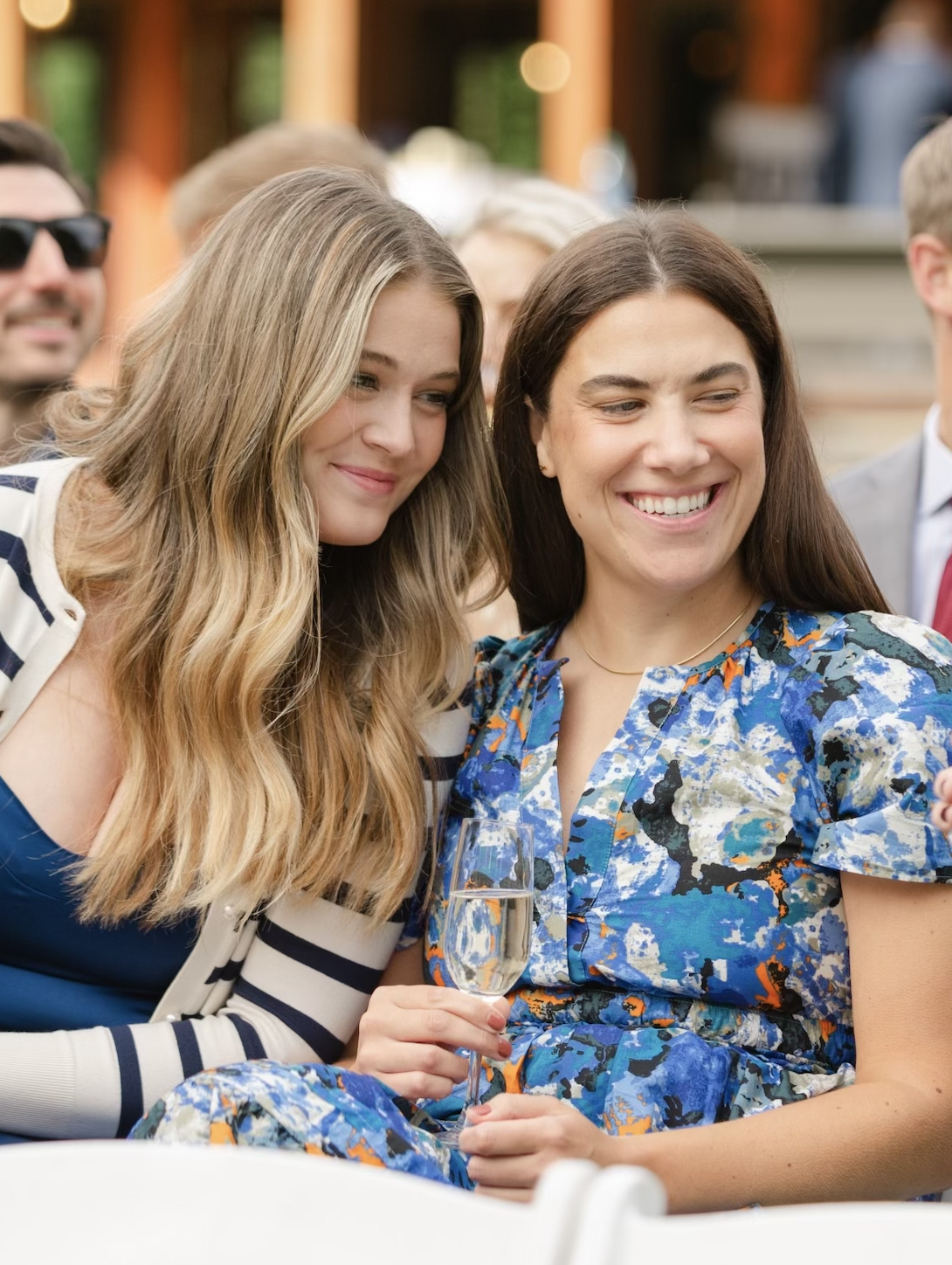 Two women are sitting close together, one holding a glass of champagne, and they are smiling at something off-camera during a social event.