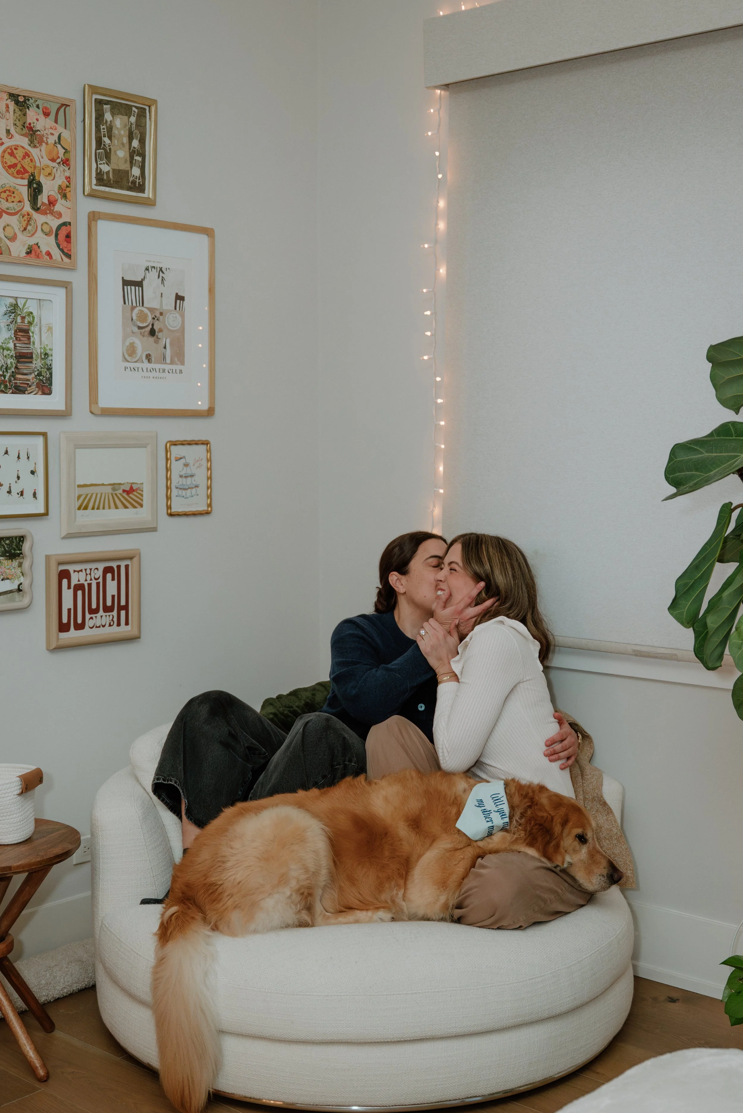 Two women sitting on a white round sofa, kissing, with a large retriever dog lying on their laps, in a cozy living room with artwork on the wall, fairy lights, a plant, and a small side table.