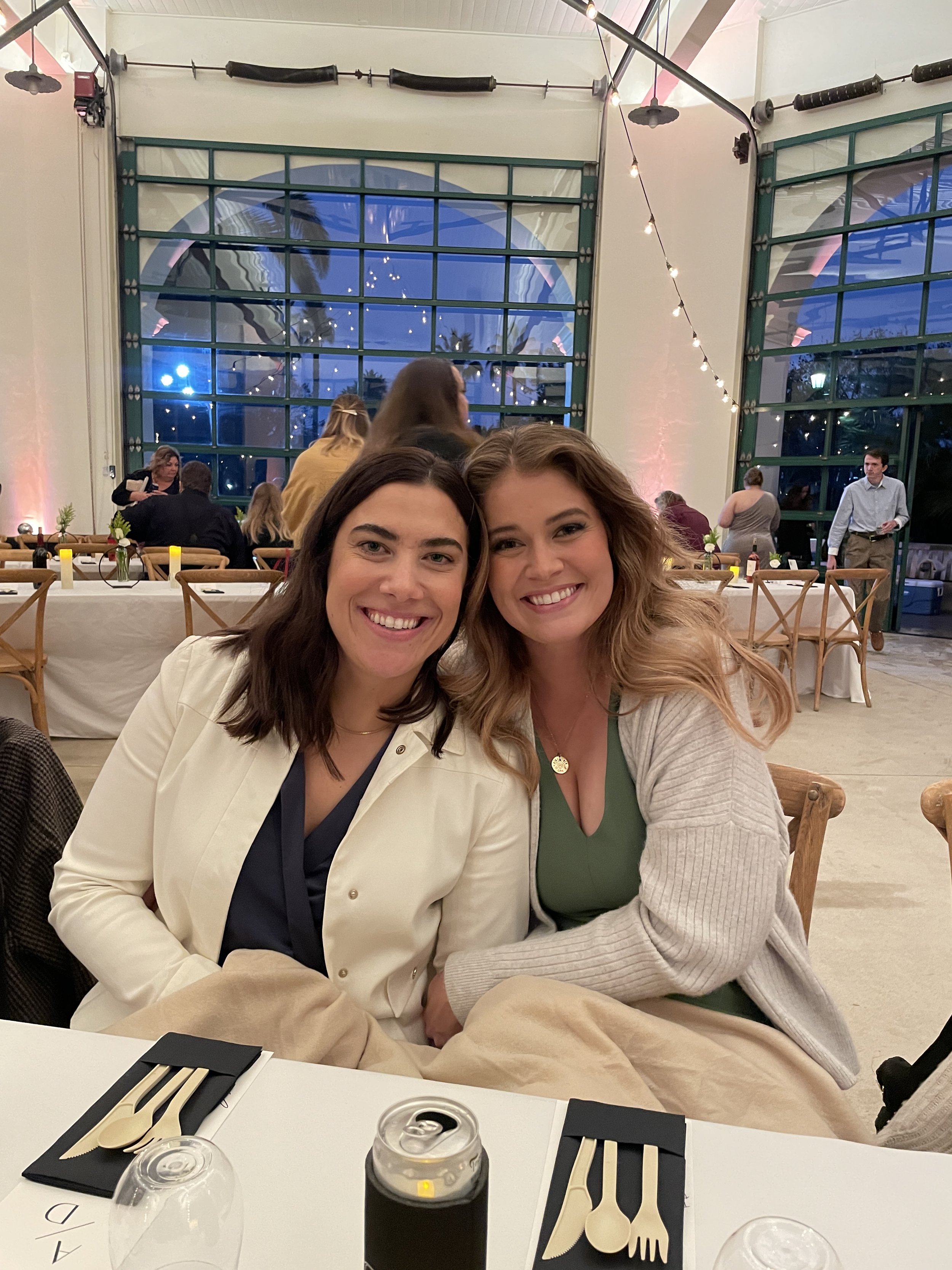 Two women sitting close together at a restaurant table, smiling and posing for the camera. The table has black napkins with beige cutlery, and glasses are visible in the foreground. The background shows other diners and large windows with a view of p