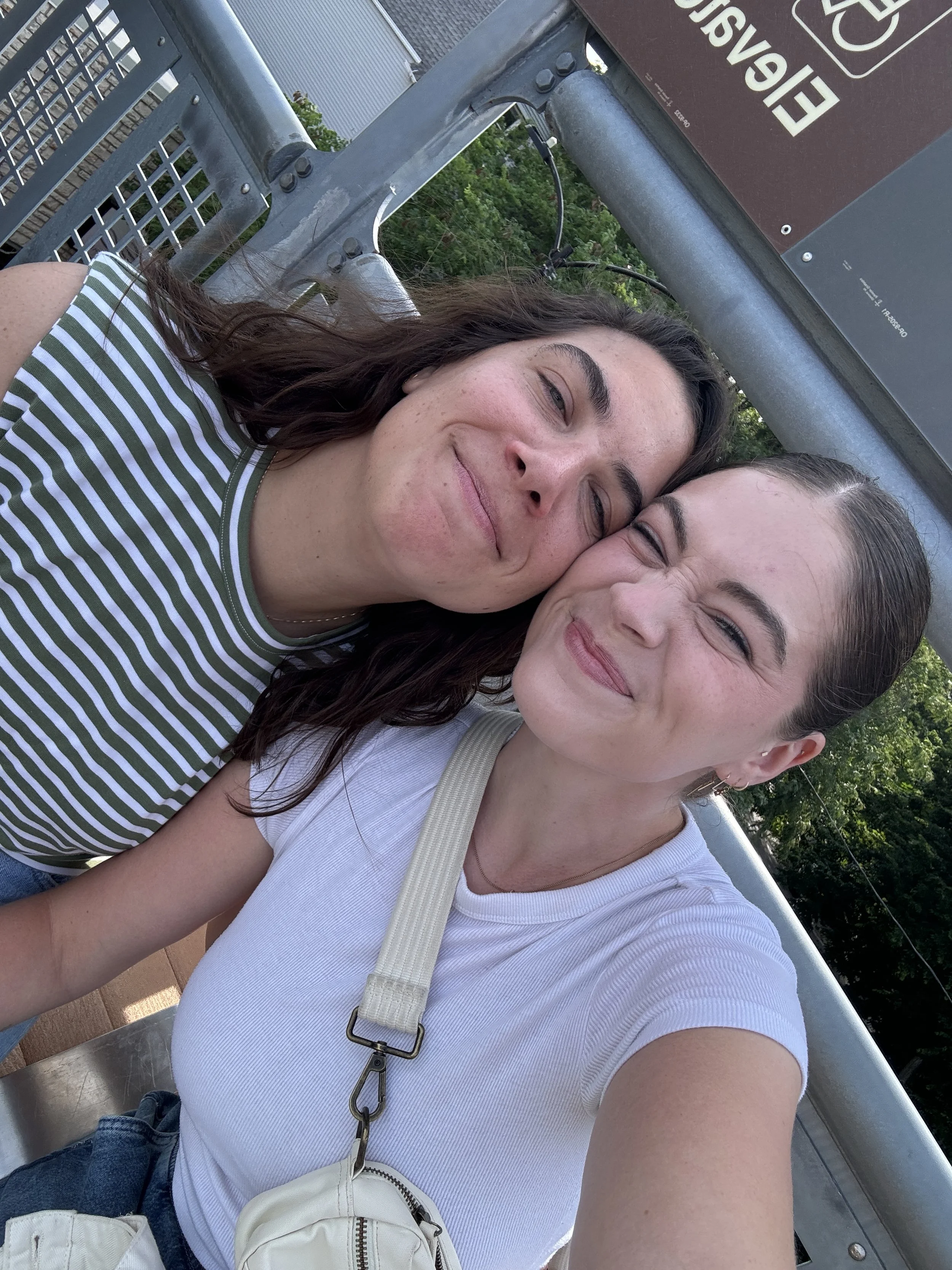 Two women smiling and leaning their foreheads together on an outdoor metal bench, with greenery in the background.