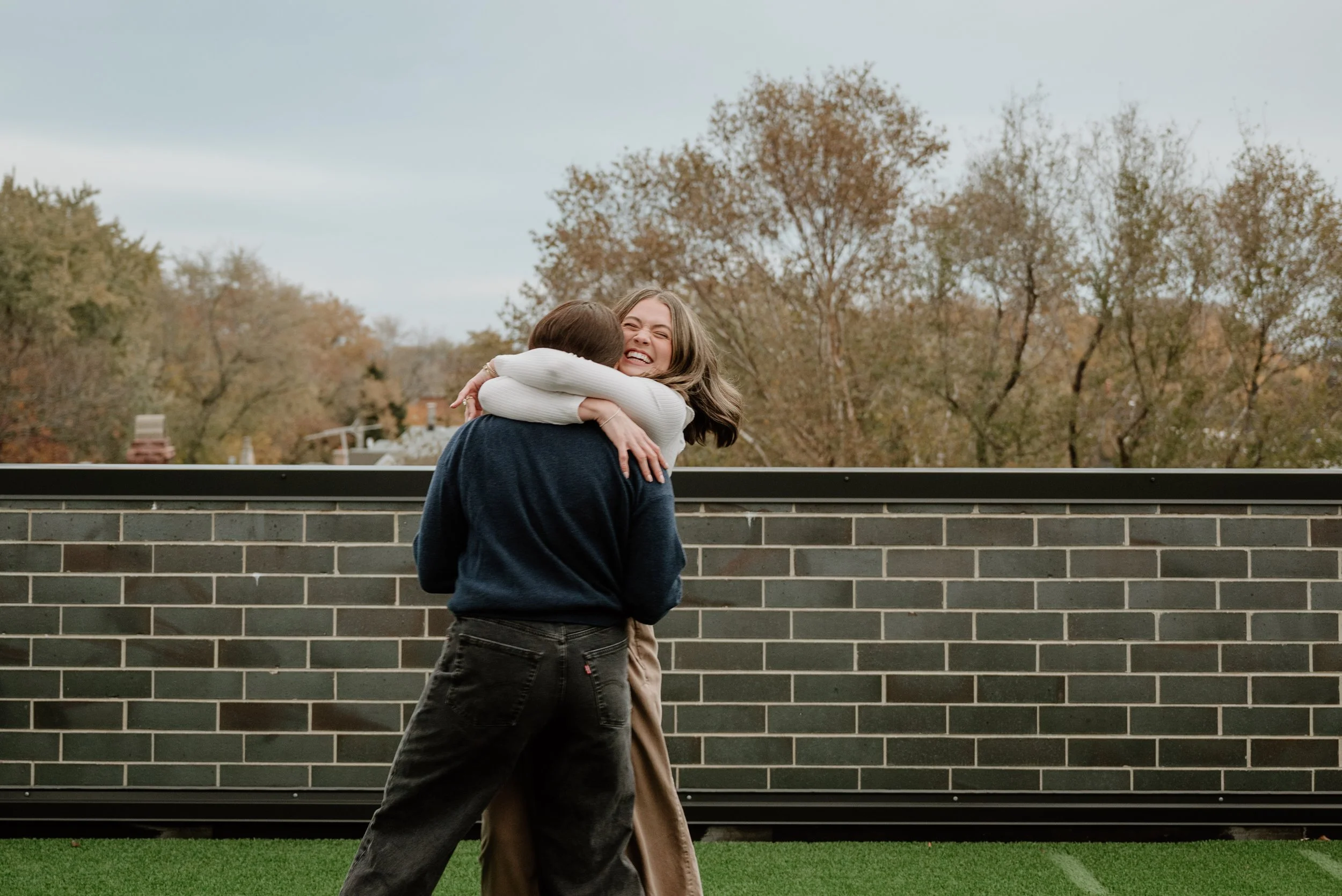 Two women hugging outdoors in front of a brick wall, with trees and a cloudy sky in the background.