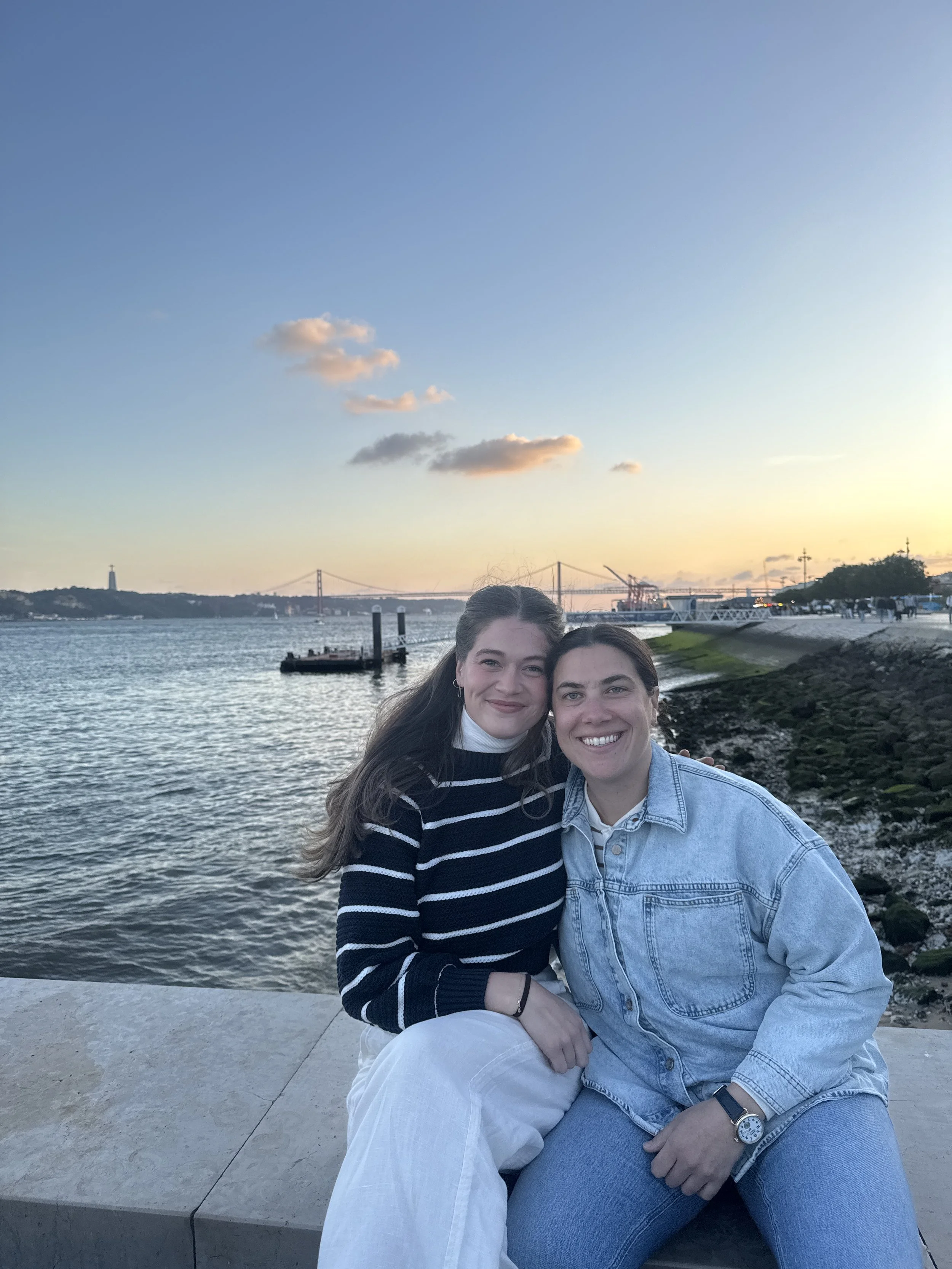 Two women sitting on a waterfront with a bridge, water, and a cityscape in the background during sunset.
