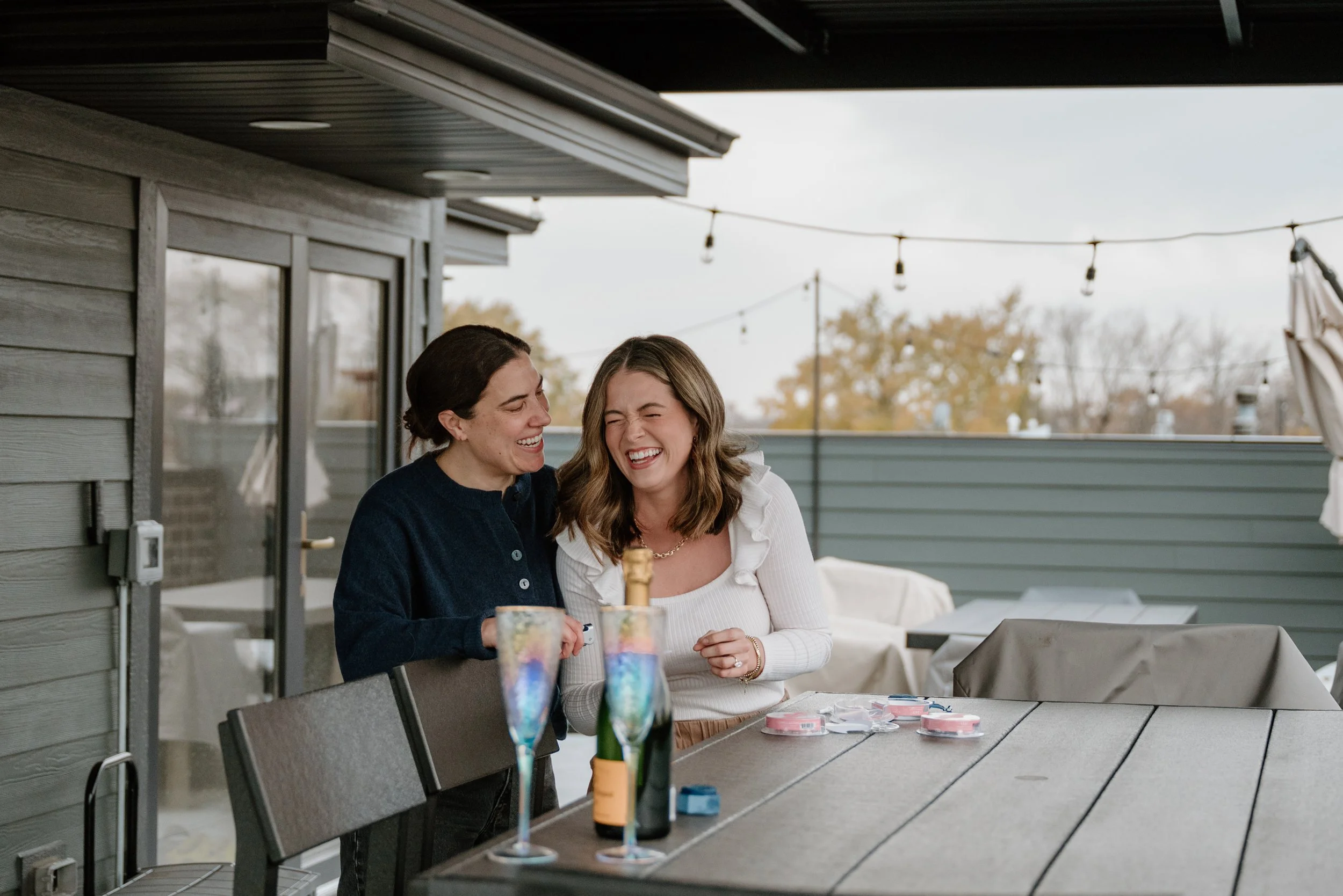 Two women laughing and smiling at an outdoor gathering on a balcony, with glasses and poker chips on the table in front of them.