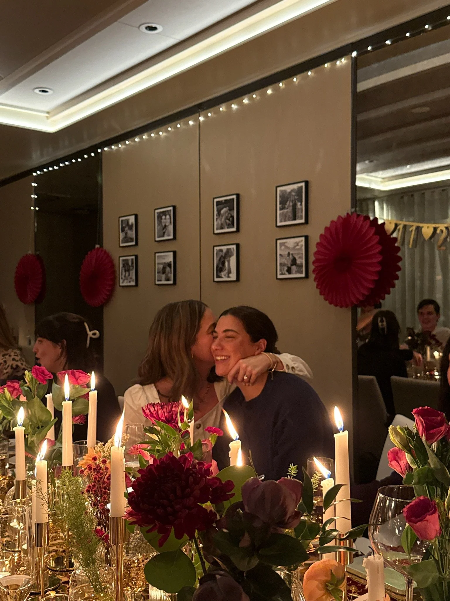 A woman kisses a woman smiling at a dinner table decorated with candles and flowers during a celebration in an indoor venue.