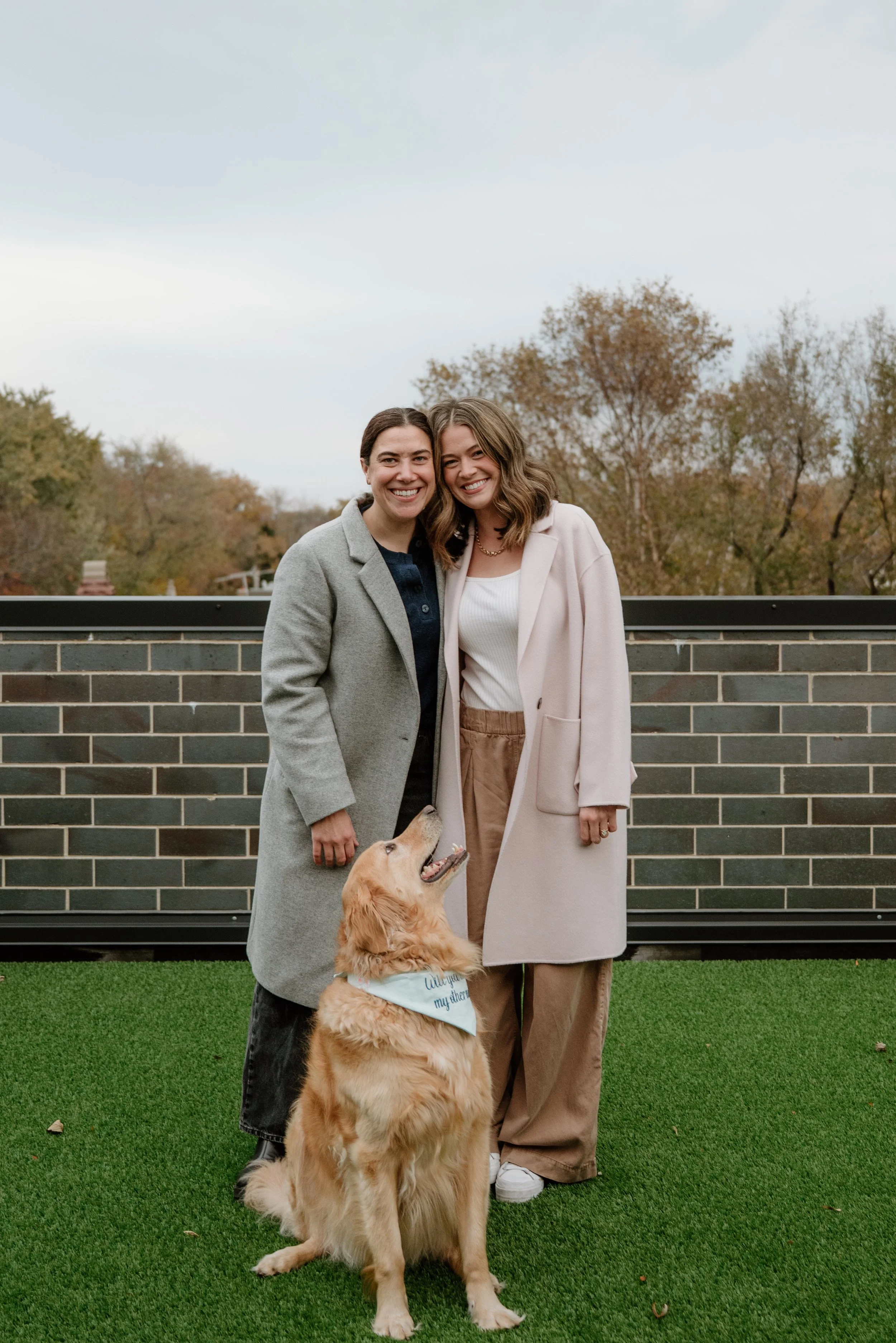 Two women smiling and standing close together outside on a rooftop, with a Golden Retriever dog sitting in front of them. The women wear casual coats, and the dog has a bandana that says 'Thank you my therapy dog.' There are trees with fall foliage i