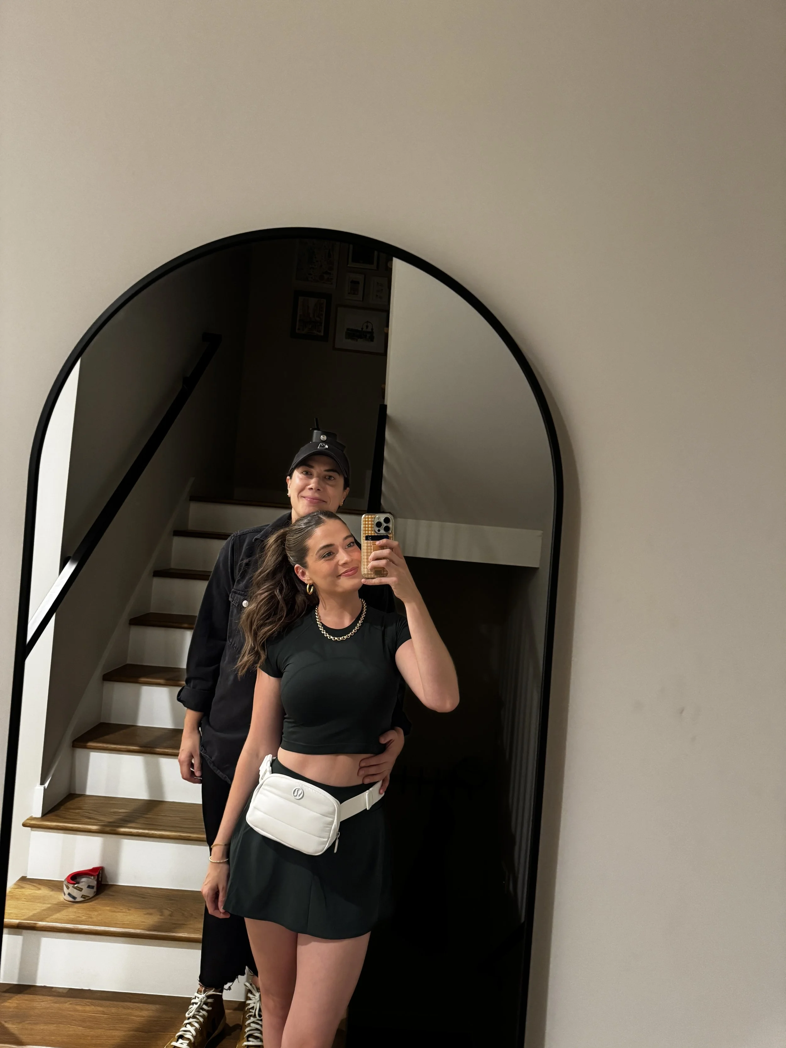Two people taking a mirror selfie in a home. The woman in the foreground has long brown hair, wears a black crop top, black skirt, and white purse. The behind her has short hair, a black cap, and black jacket. The background shows a staircase with wo