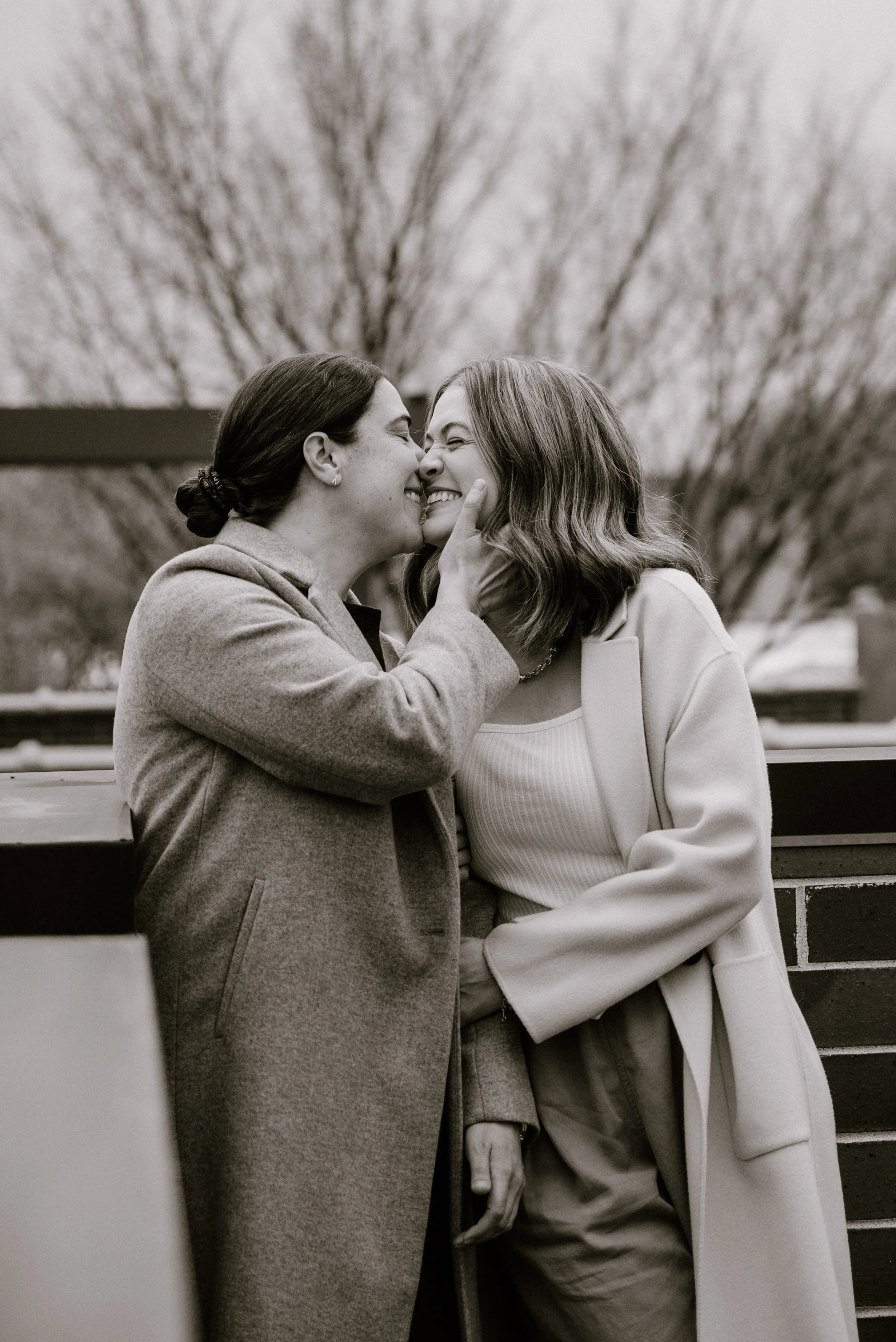 Two women share a gentle, happy moment, touching noses and smiling warmly at each other outdoors during winter, with leafless trees in the background.