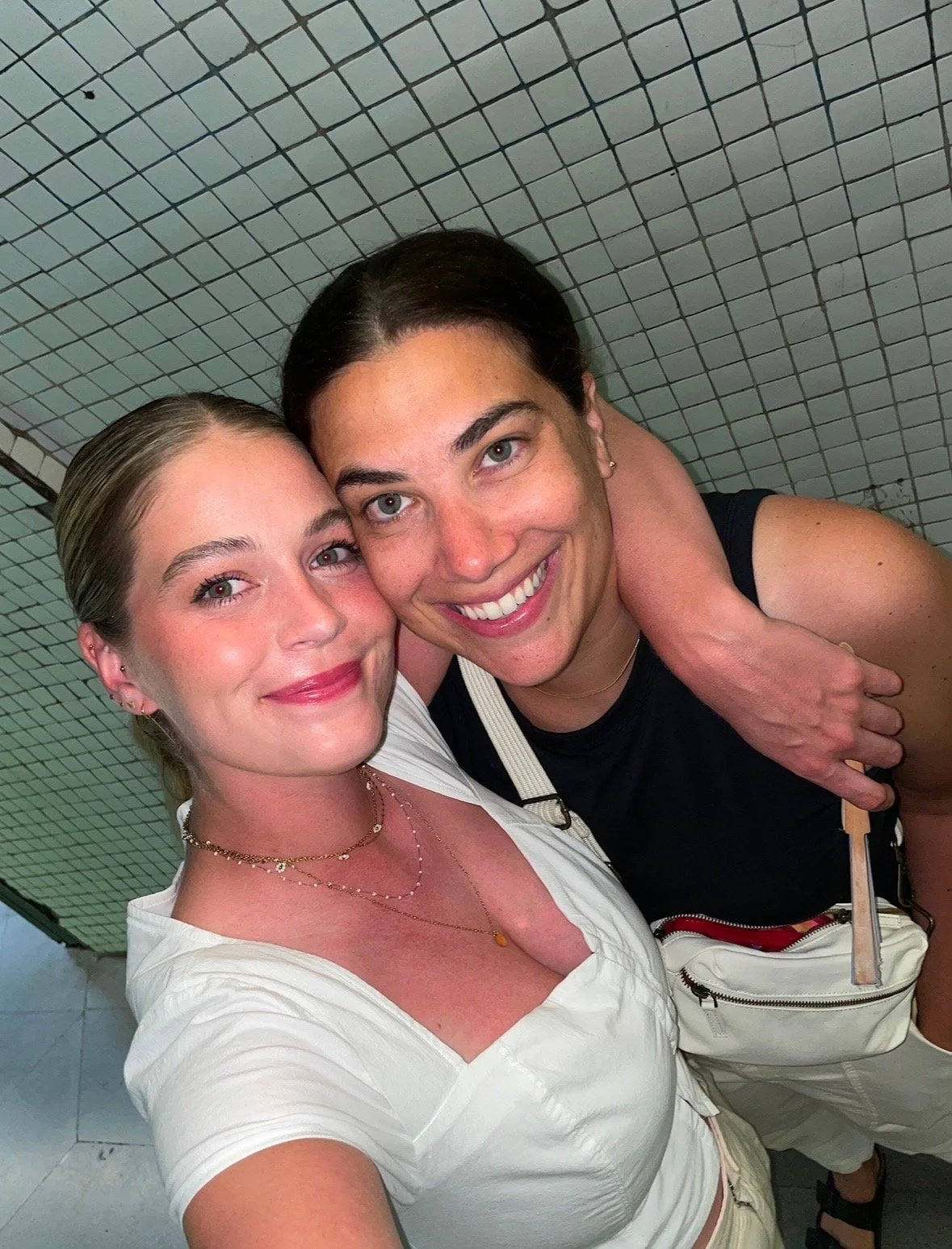 Two women taking a selfie together, smiling, with mosaic tiled wall in the background. One woman has dark hair and the other has light hair, both with their arms around each other.