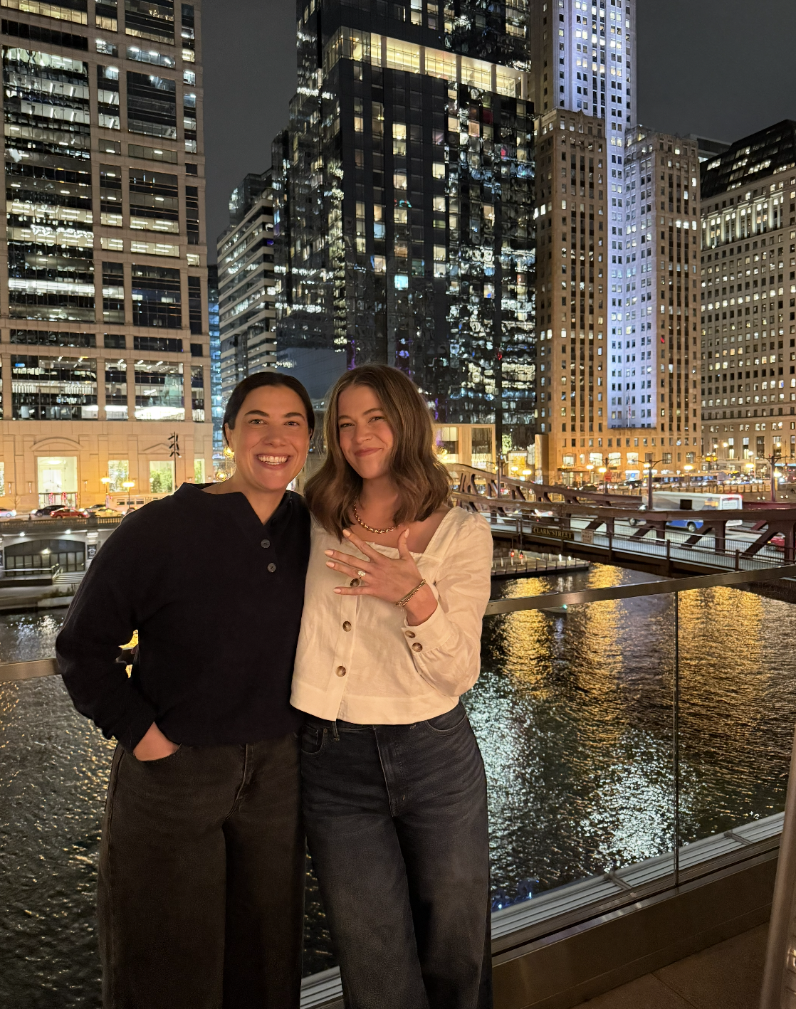 Two women standing together at night in an urban setting with tall illuminated buildings, a bridge, and water reflecting city lights in the background. One woman is smiling with her hand on her chest, and the other is smiling with her hand in her poc