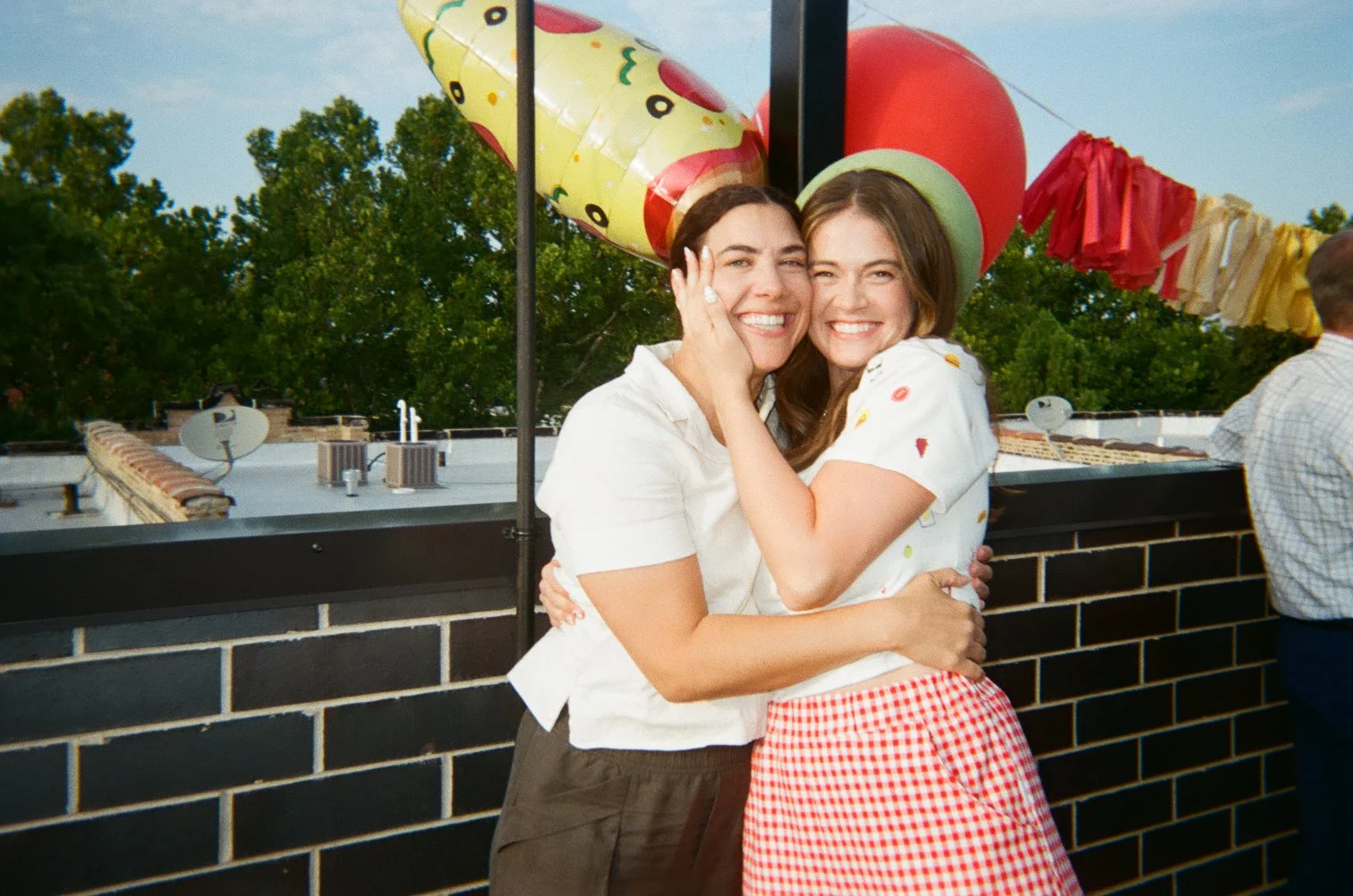 Two women happily hugging at a birthday party outdoors, with colorful balloons and decorations in the background.