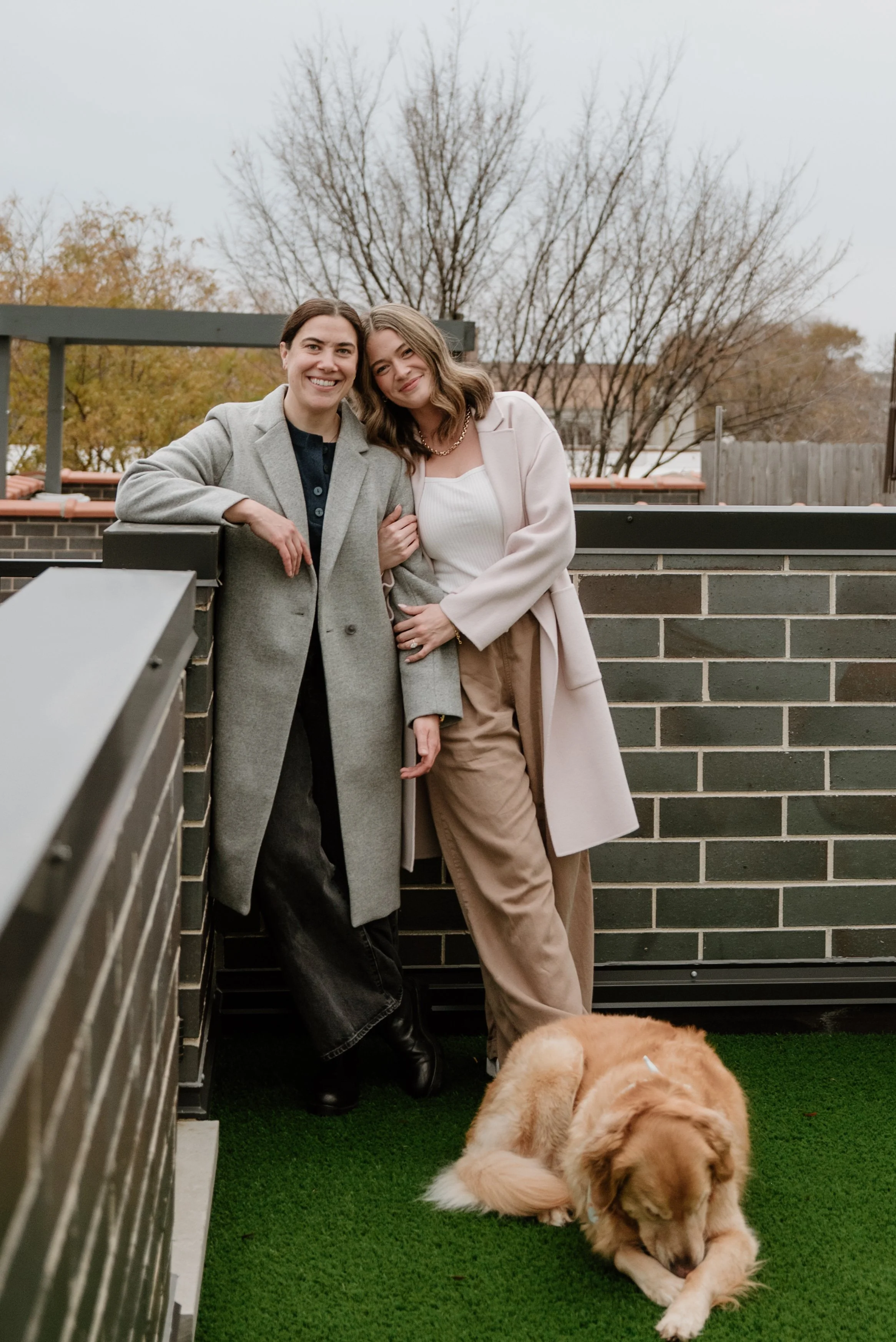 Two women stand close together on a balcony with a brick wall, wearing coats and smiling, with a large golden retriever lying on green artificial grass in front of them, and leafless trees in the background on a cloudy day.