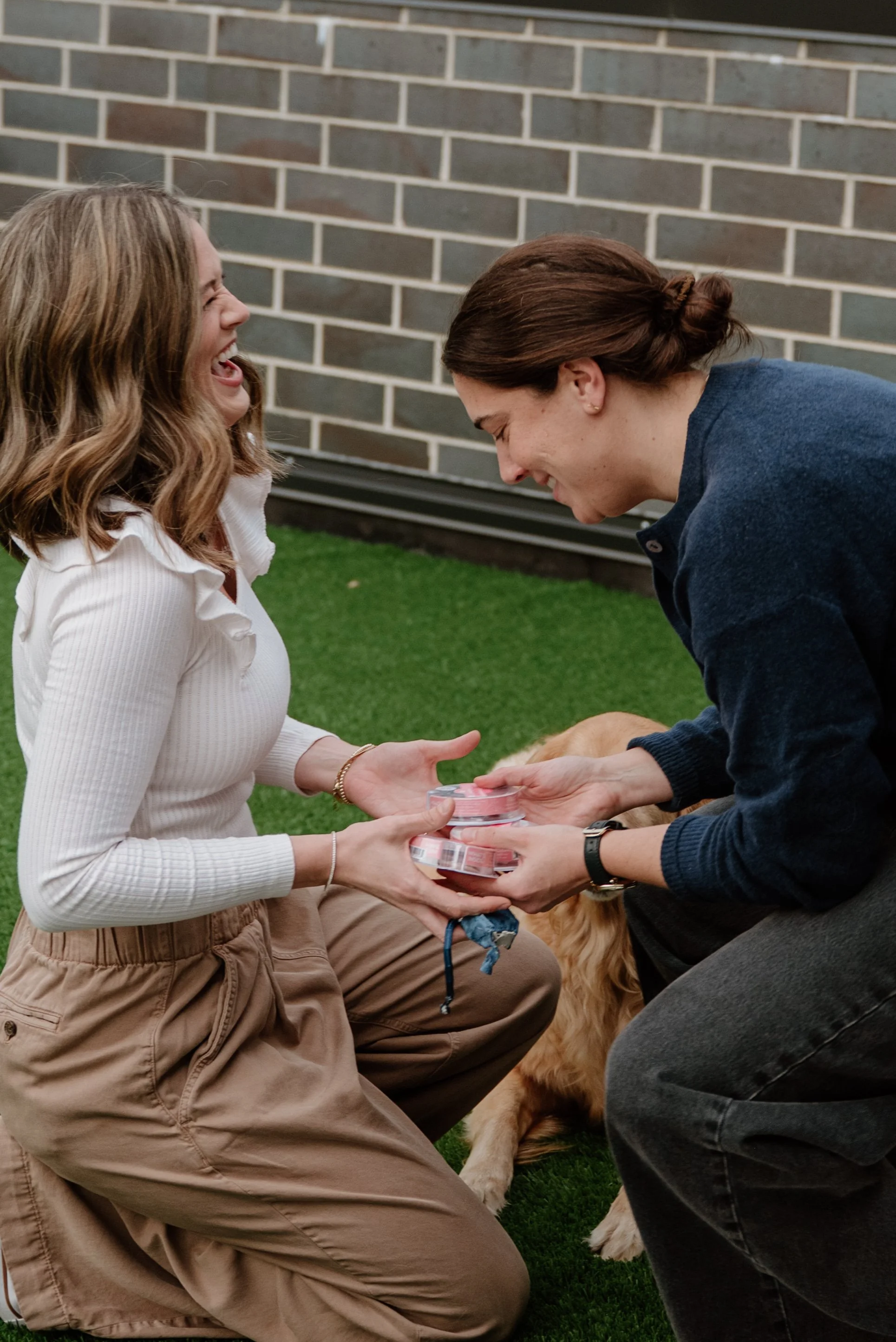 Two women sharing a moment while kneeling on a grassy area outdoors in front of a brick wall, holding small containers, with a dog partially visible at the bottom of the frame.