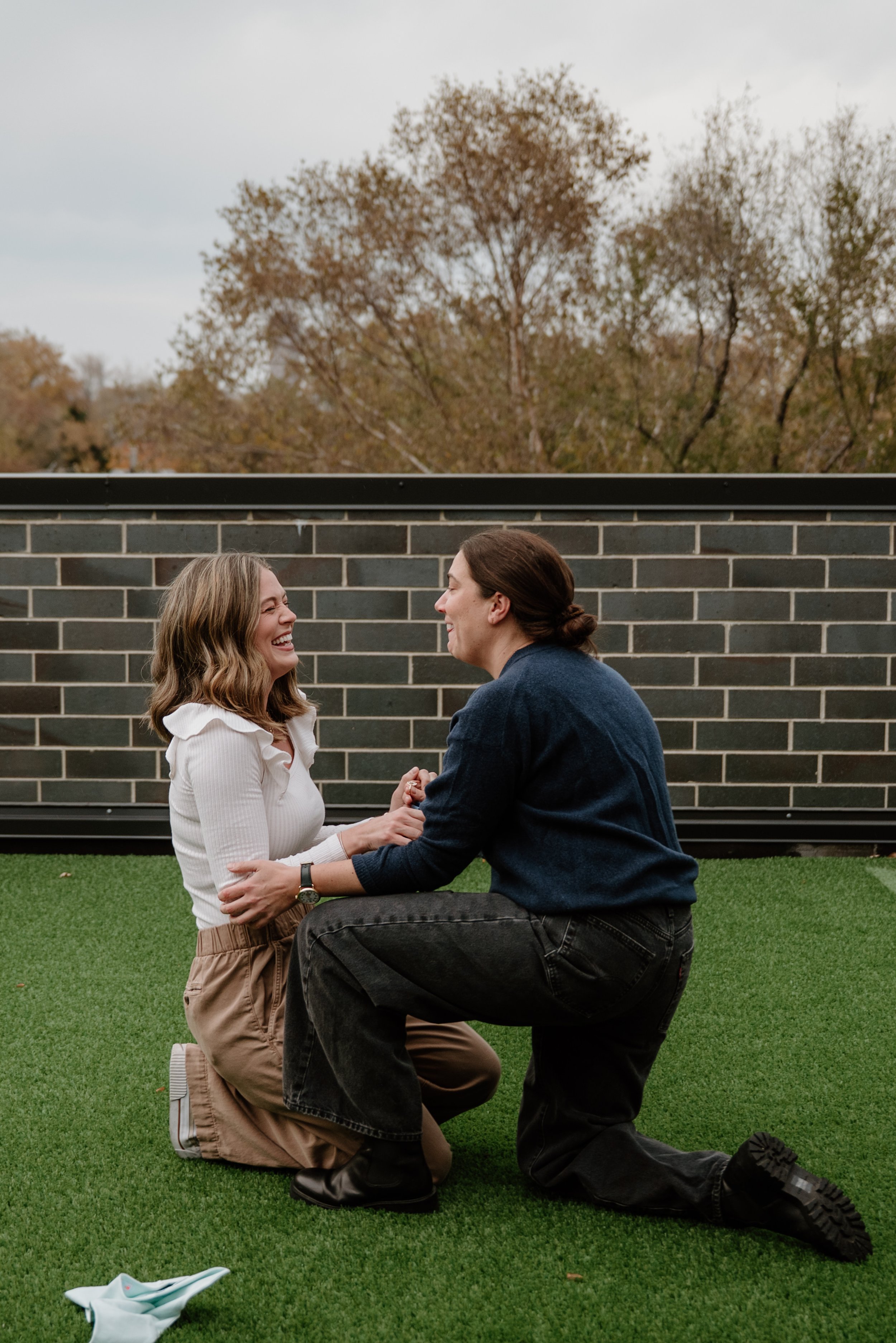 A woman kneeling on one knee holding hands with a woman standing opposite her, both smiling and looking at each other outdoors on a green lawn with a brick wall and trees in the background.