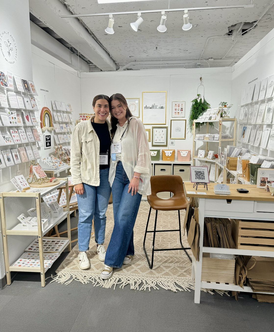 Two women smiling and standing close together inside a gift and stationery store, surrounded by greeting cards, framed artwork, and decorative items.