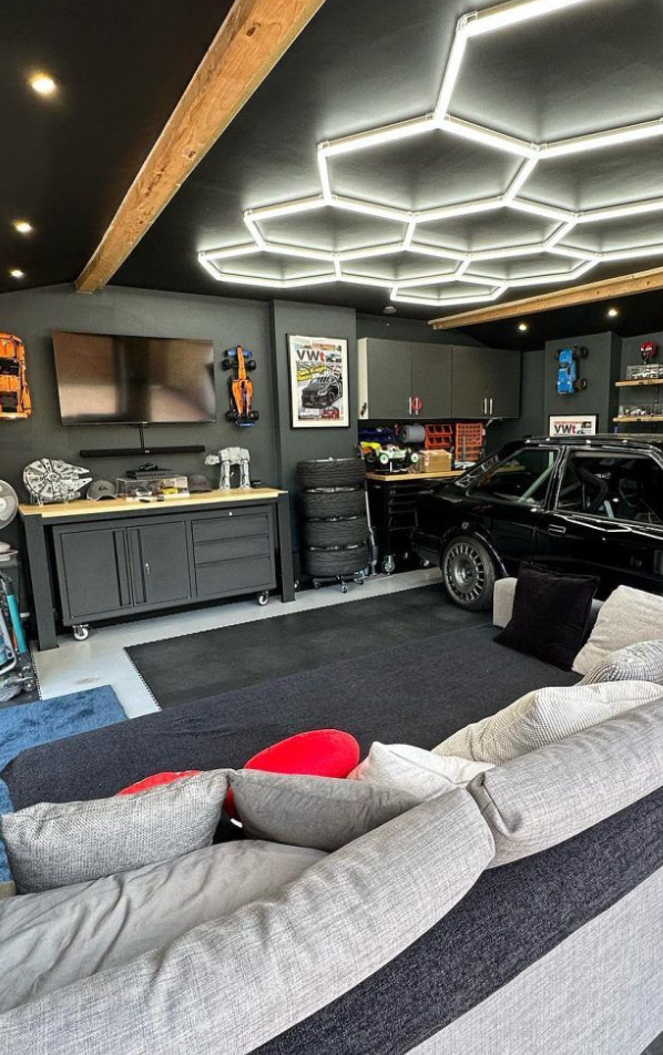 A modern garage with a black sports car, gray cabinets, a flat-screen TV, tires, and garage tools. A sectional sofa with pillows is in the foreground, and a unique hexagonal LED ceiling light fixture.