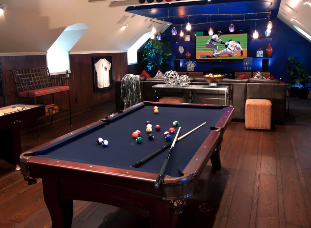 A game room with a pool table in the foreground, a television displaying a baseball game, and a comfortable seating area with cushions in the background.