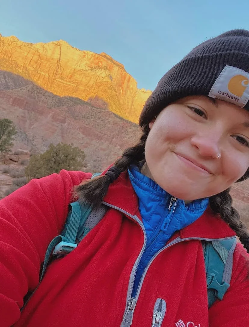 Della is wearing a beanie and smiling while outdoors.