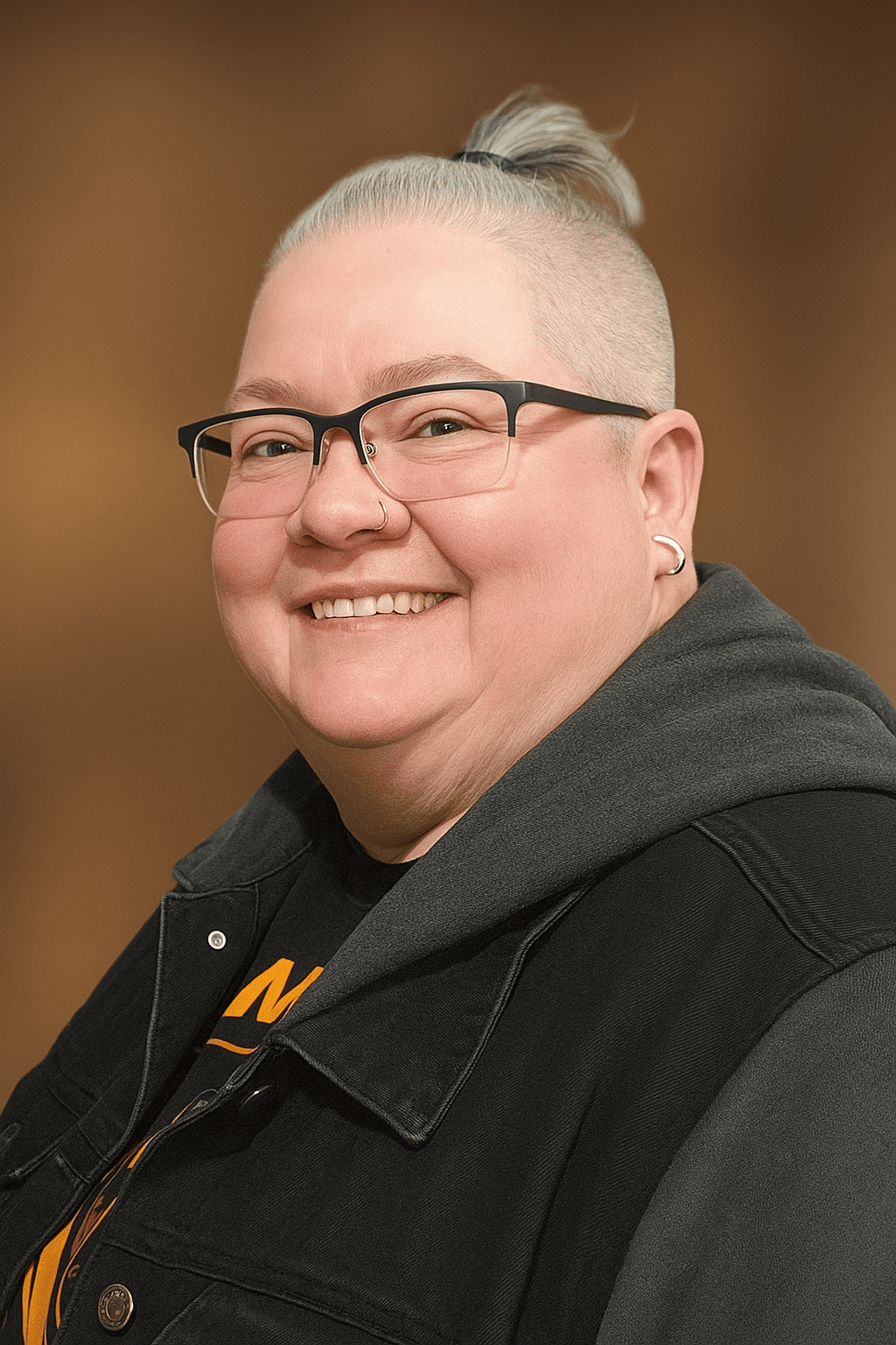 Smiling woman with light blonde hair in a top knot, wearing glasses, earrings, a nose ring, a black denim jacket, and a black shirt with orange text, standing against a brown background.