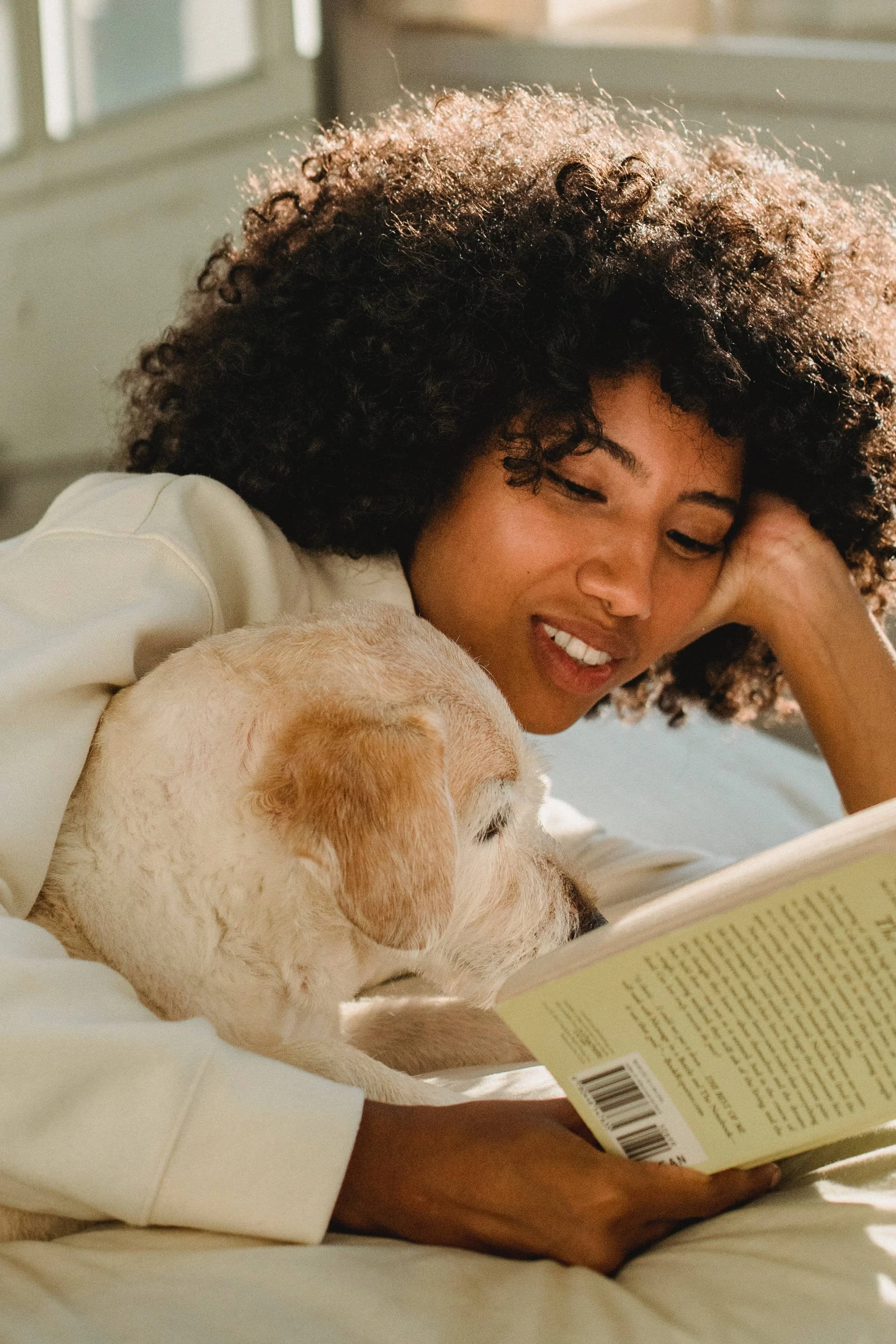 A person with curly hair is lying down reading a book with a light-colored dog.
