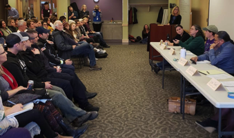 A group of people sits in rows of chairs facing a panel of four individuals seated at a long table. 