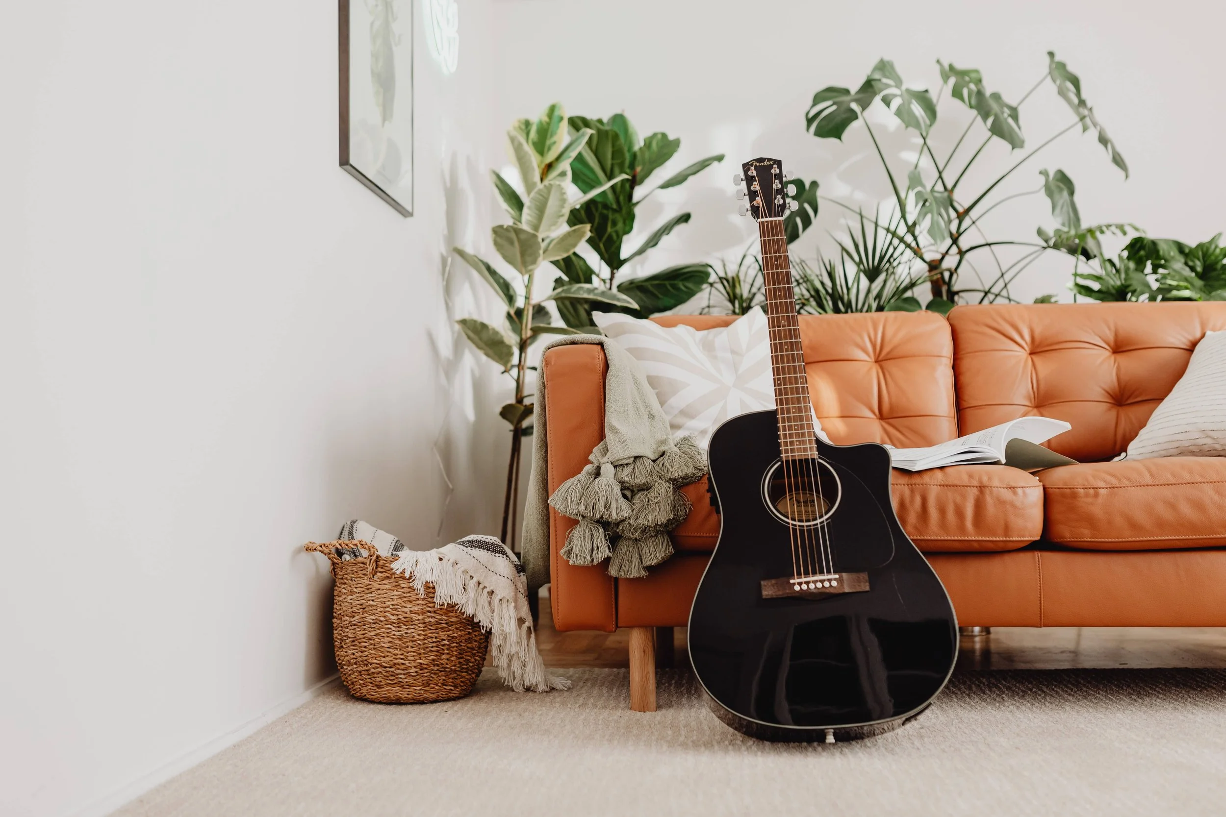 A black acoustic guitar leaning against an orange leather sofa in a living room with potted plants, a basket with a throw blanket, and an open book on the sofa.