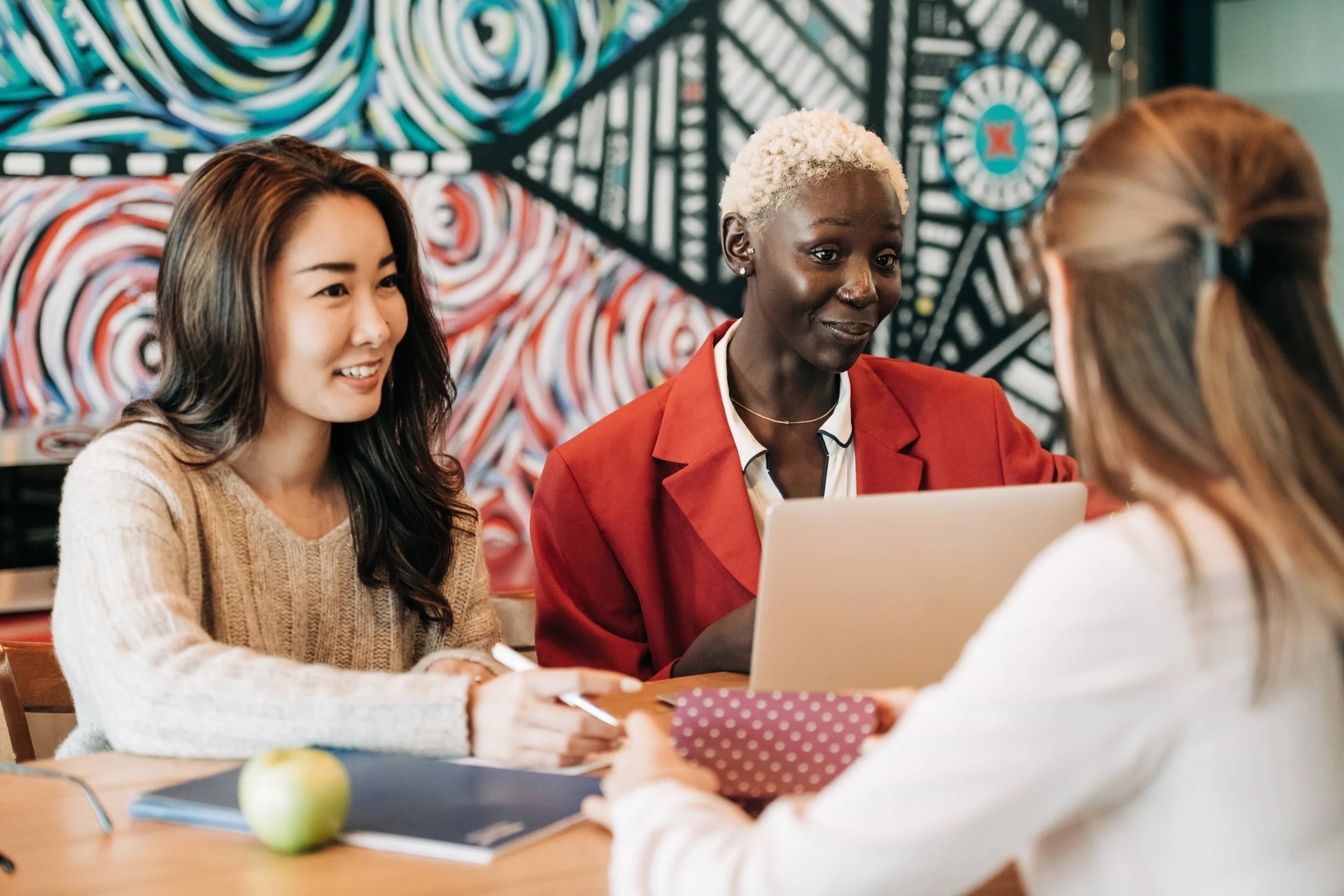 Three women having a meeting at a table in a cafe with a colorful, abstract mural in the background. One woman, with curly dark hair in a beige sweater, is smiling and holding a pen. The second woman, with short, bleached hair and wearing a red blazer, is looking at a laptop. The third woman, with long, light brown hair tied back, is facing away from the camera and holding a folder or notebook.