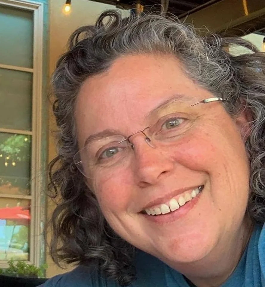 Smiling person with curly hair and glasses, seen in a close-up portrait.