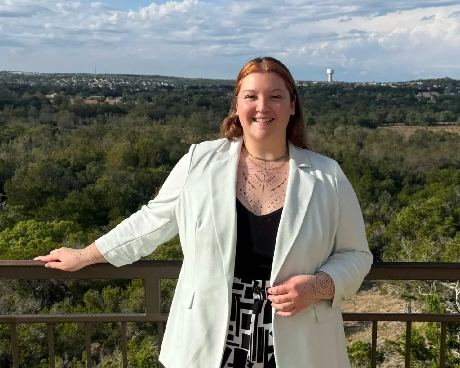Stacy (they/she) is smiling, standing outdoors on a balcony with a scenic view of trees and distant landscape under a partly cloudy sky.