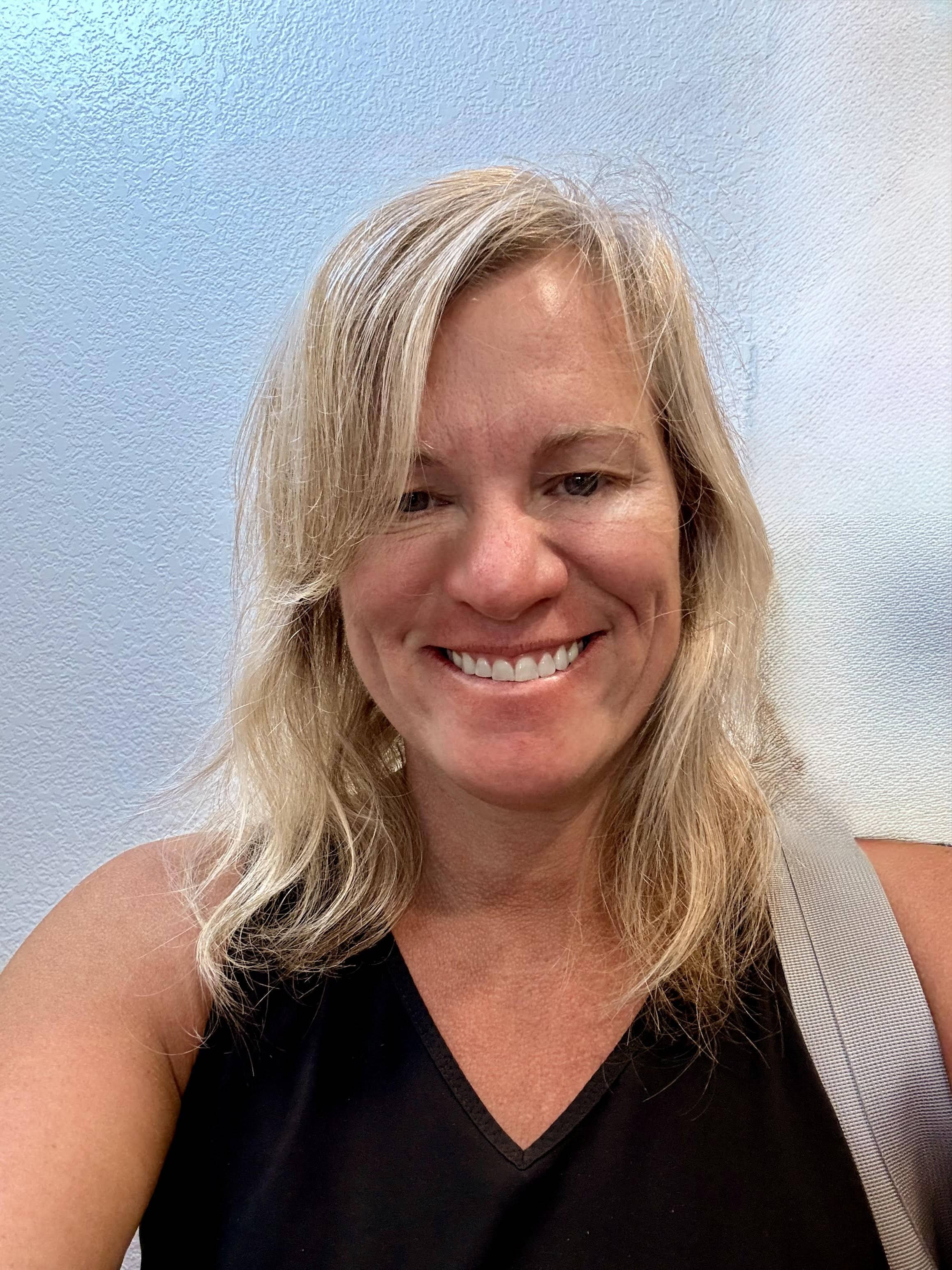 A woman with shoulder-length blonde hair, smiling, wearing a black sleeveless top and a gray strap, standing against a light blue textured wall.