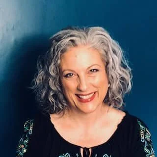 A person with curly gray hair smiles warmly against a dark blue background.