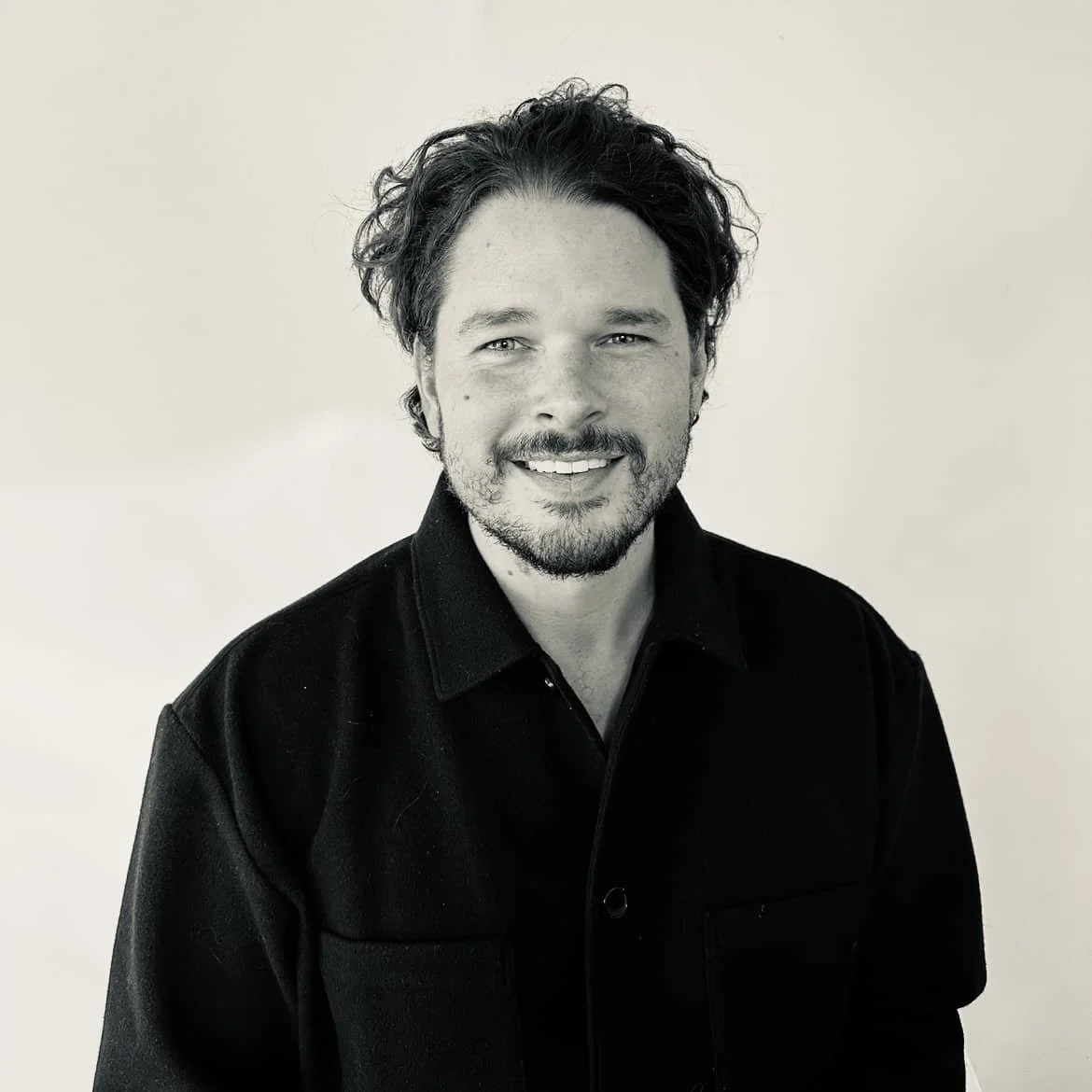 Black and white portrait of a smiling man with wavy dark hair, a beard, and a mustache, wearing a black shirt against a plain background.