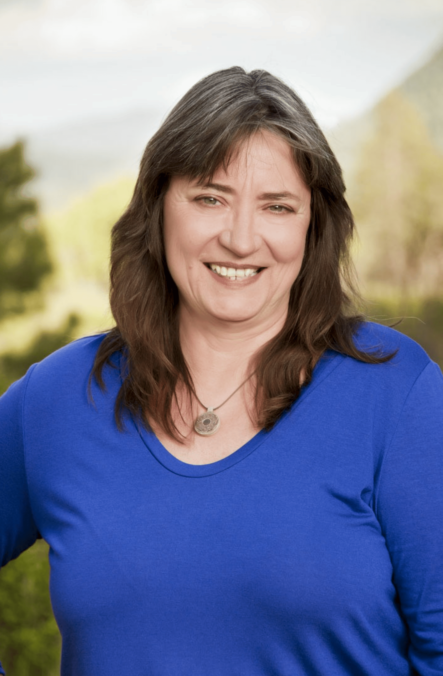 A woman with long brown hair, wearing a blue top and a silver necklace, stands outdoors with blurred green trees and mountains in the background.