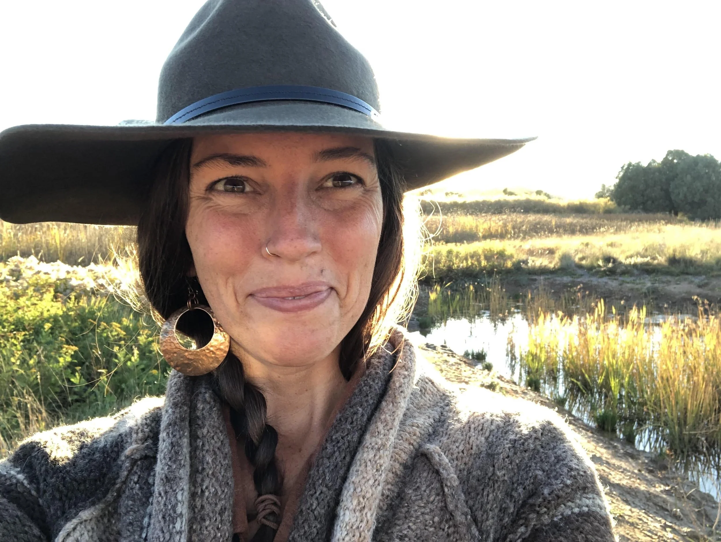 Person smiling, wearing a wide-brimmed hat and large earrings, standing outdoors.