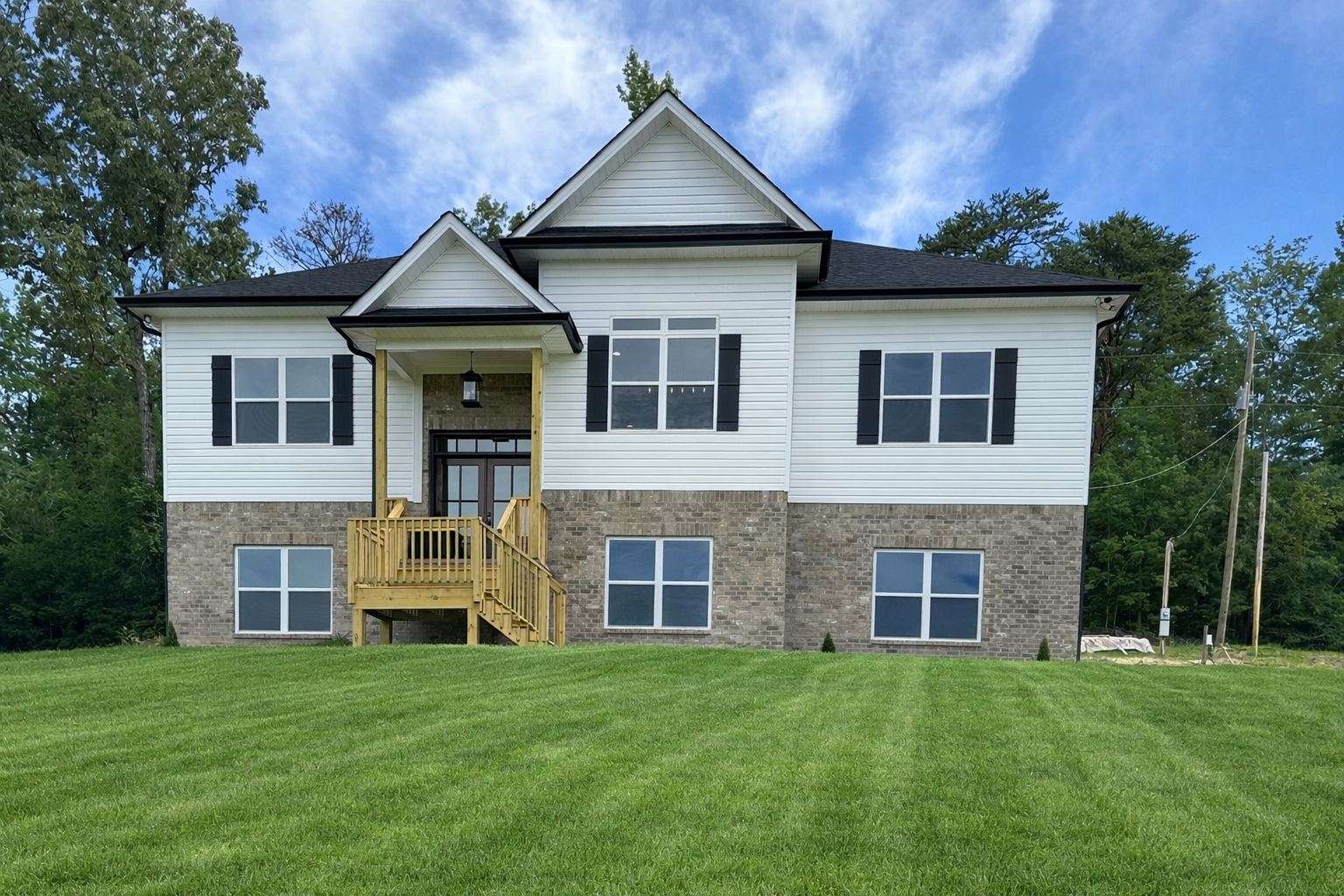 Two-story family house with white siding, brick lower level, black shutters, a small front porch with stairs, surrounded by green grass and trees, under a partly cloudy sky.